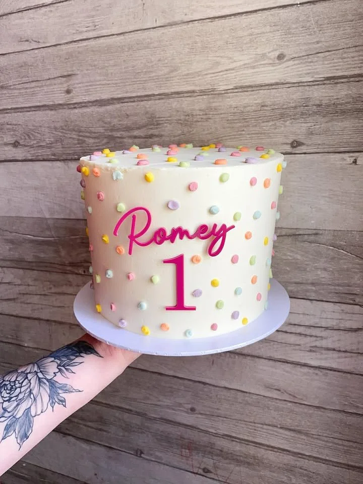 A white cake decorated with colorful round sprinkles, with pink text that reads "Roney" and a large pink number "1" for a first birthday celebration.