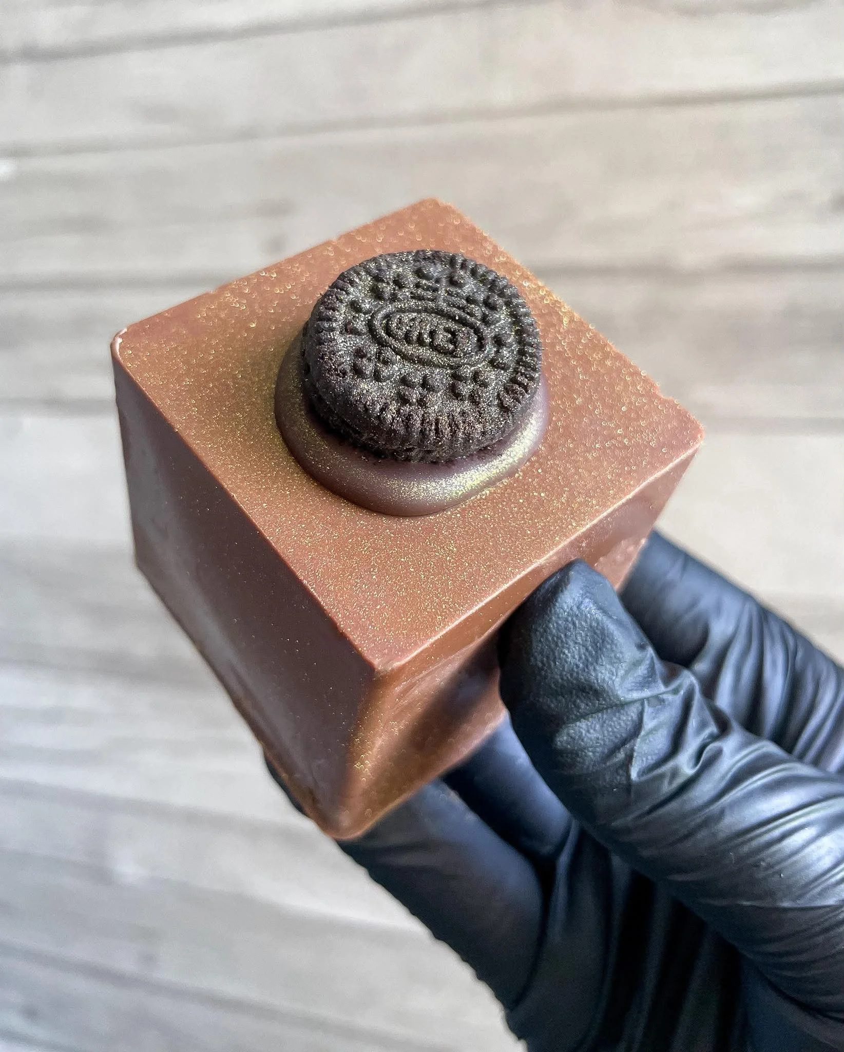 Close-up of a small, round Oreo cookie on a pink, glittery box, held by a person wearing a black glove, with a white background.