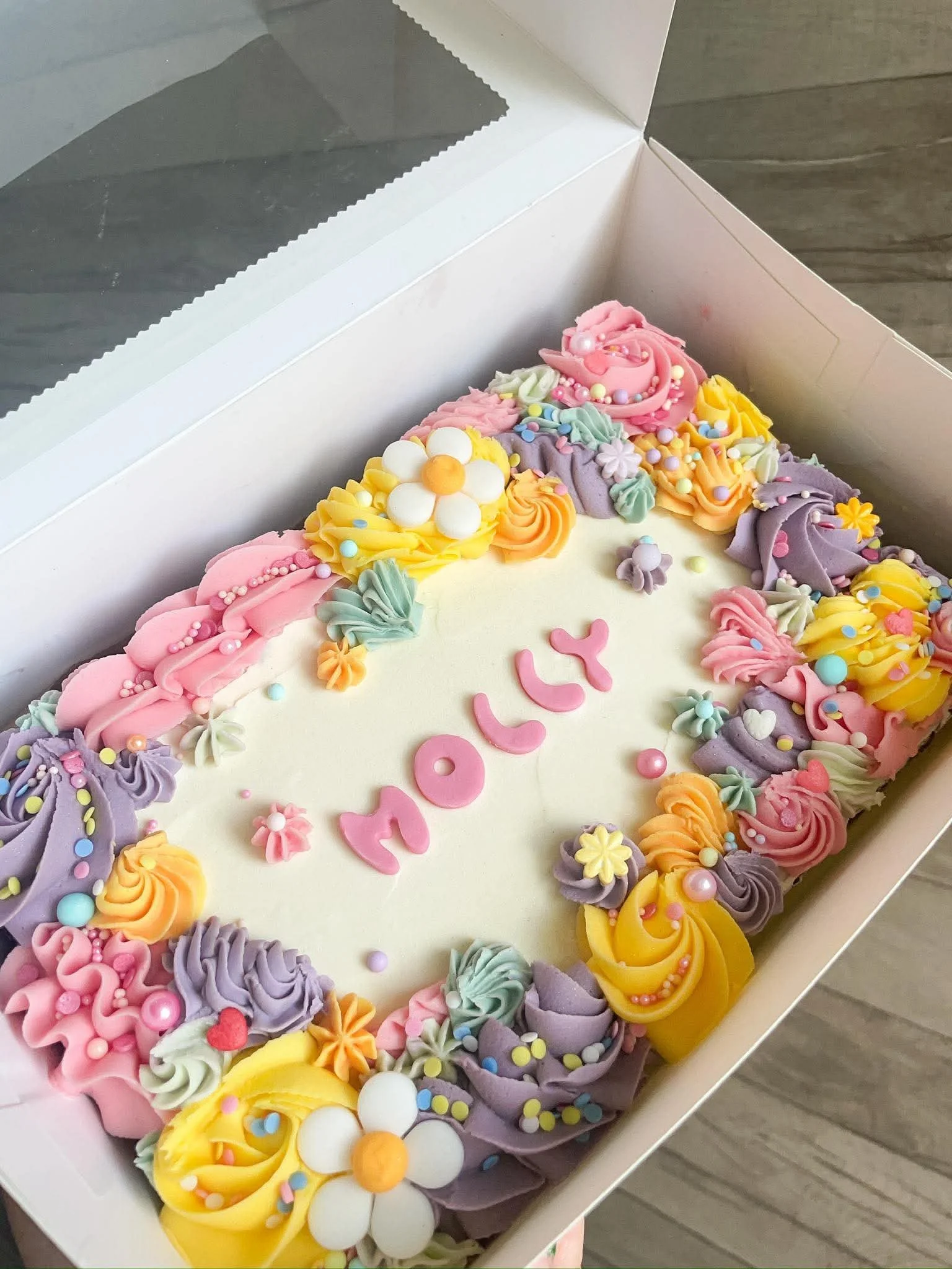 Colorful decorated birthday cake with pink, yellow, purple, and white frosting, topped with various small decorative candies and flowers, in a white box.