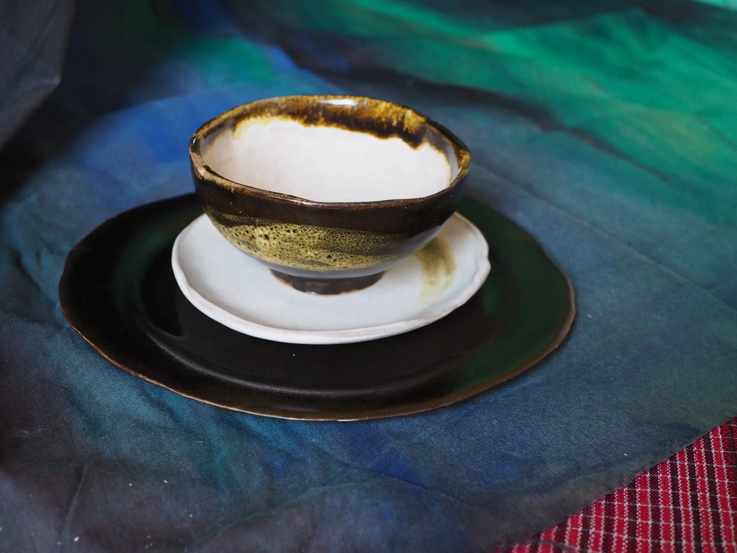 A handcrafted ceramic bowl with dark brown and beige glaze, sitting on a white saucer, on top of a black tray. The tray is on a dark, textured fabric with a multicolored appearance, next to a red and black checkered cloth.