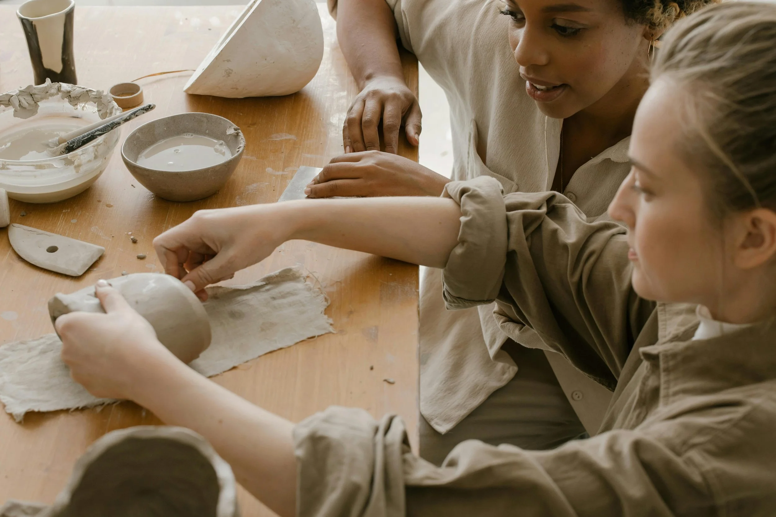 Two women and a young girl baking pottery together at a wooden table. The table has bowls, clay, and sculpting tools.