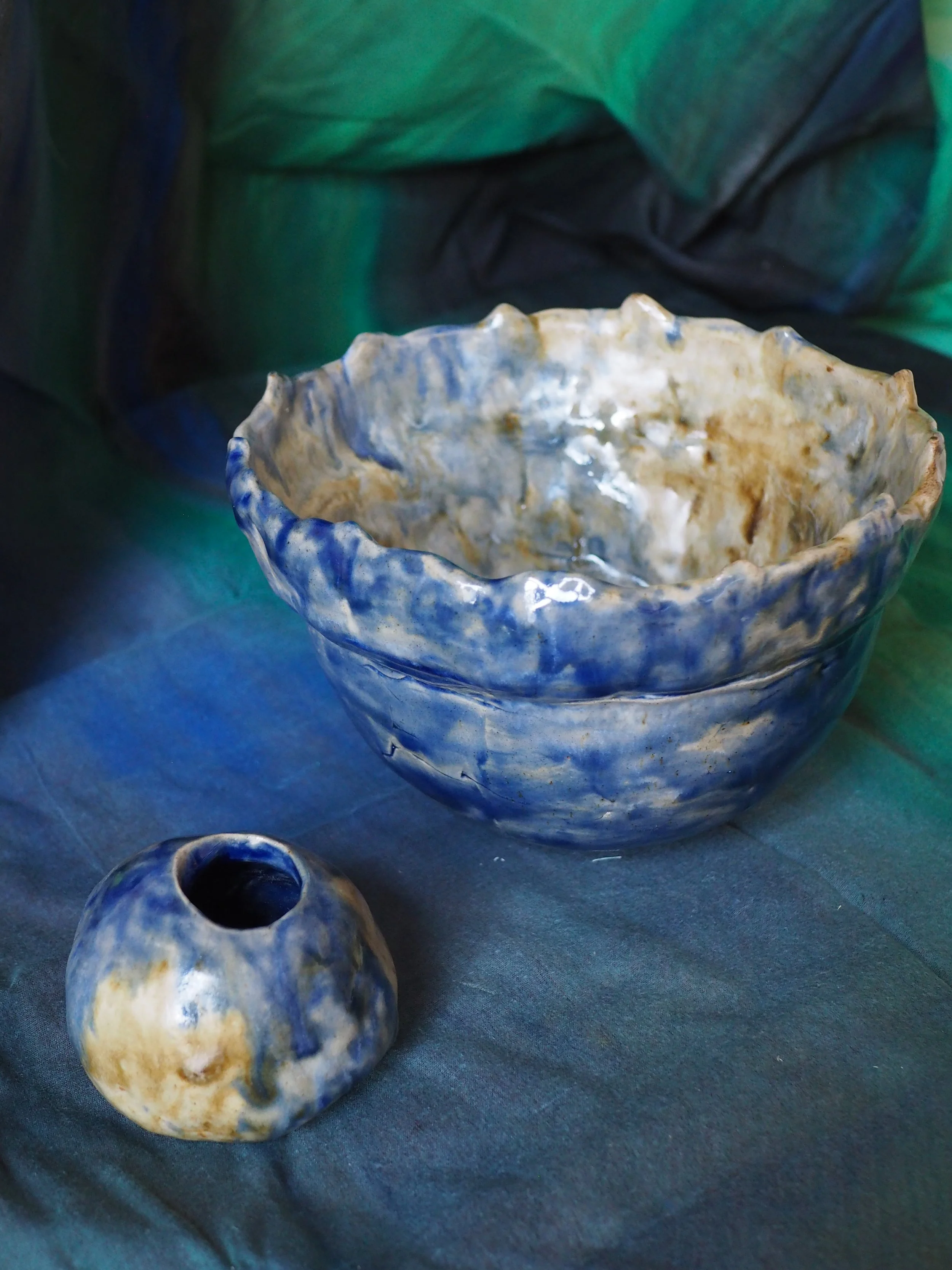 Two handmade ceramic bowls, one larger with a wavy rim and blue and beige glaze, and a smaller one with a similar glaze, on a dark fabric surface with a background of green and black fabric.