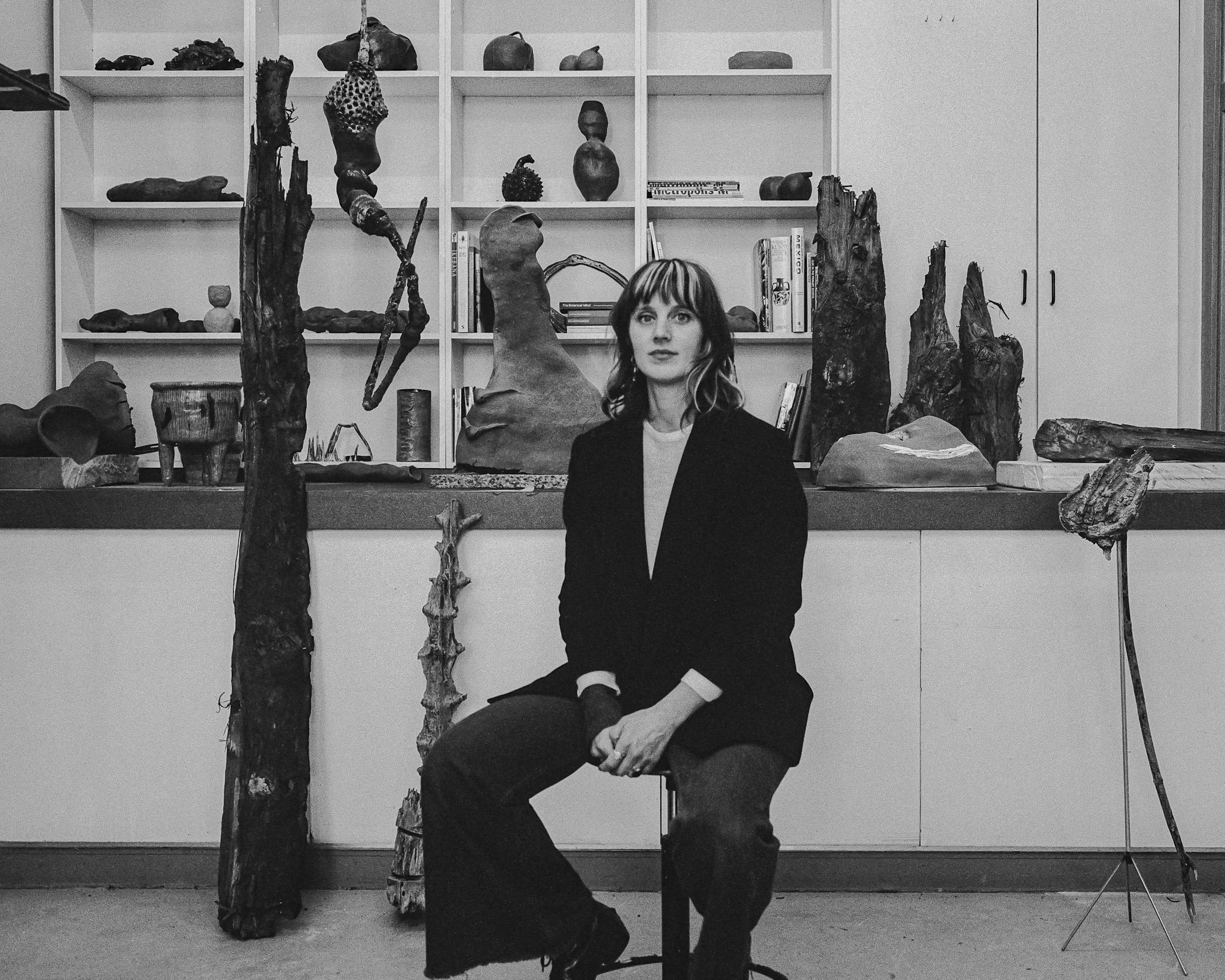 A woman with shoulder-length hair, dressed in a dark blazer, sitting in front of a display of various sculptures and artifacts in a room with bookshelves, modern art pieces, and a cabinet.