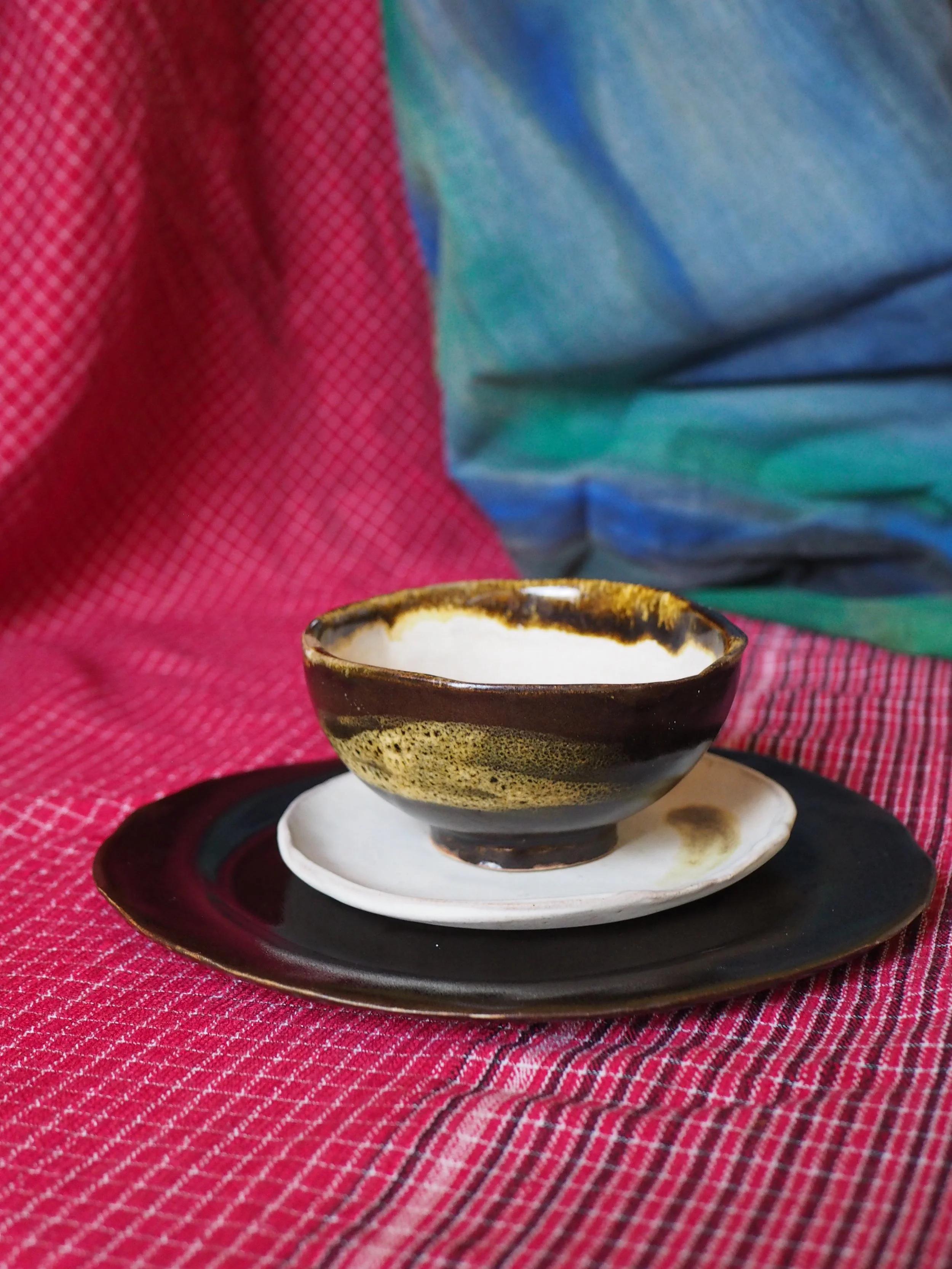 A ceramic bowl with a brown and beige glaze on a white saucer, set on a black platter, on a red and black striped tablecloth.