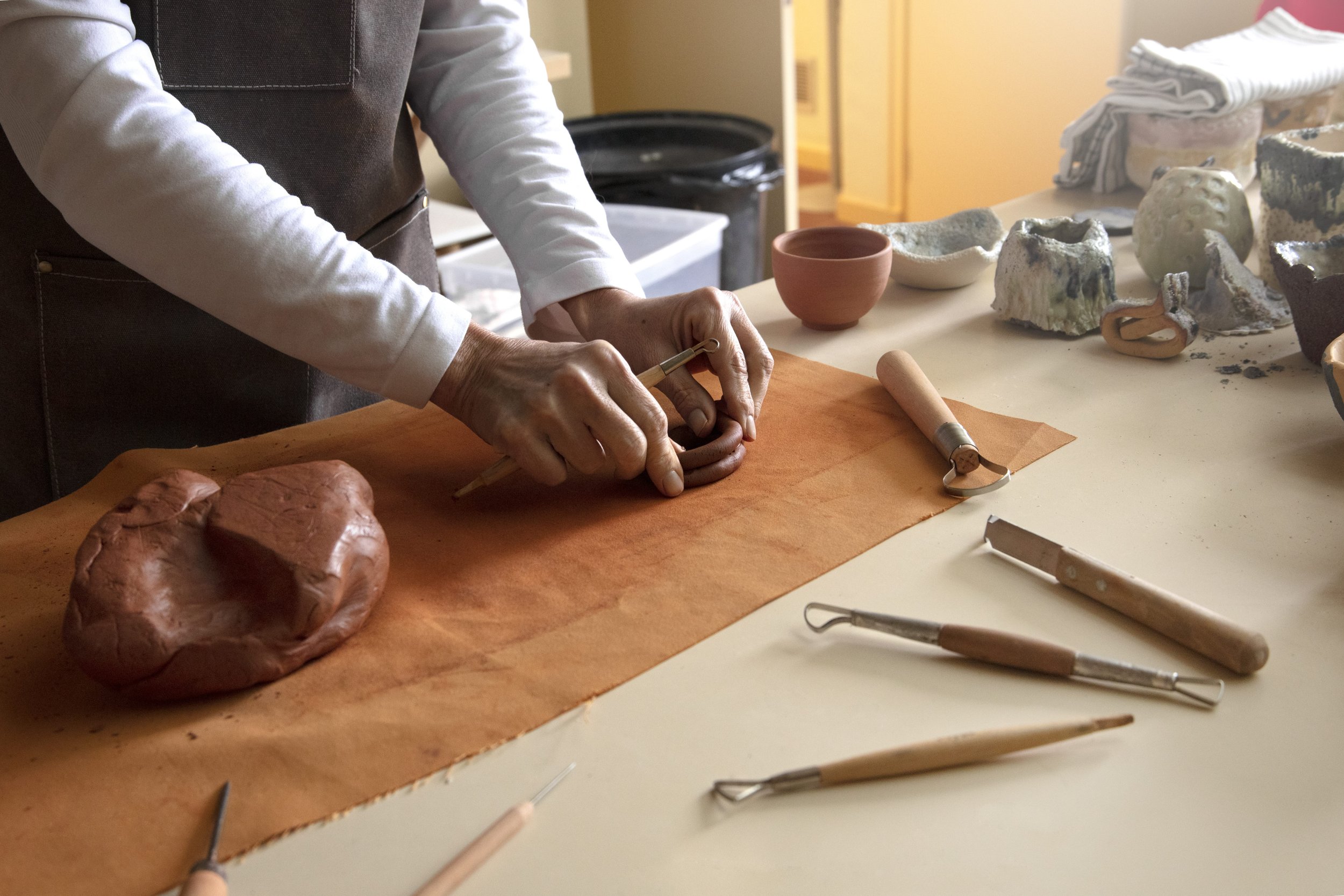 Person shaping clay on a table with pottery tools, various clay objects and ceramic pieces on the surface.
