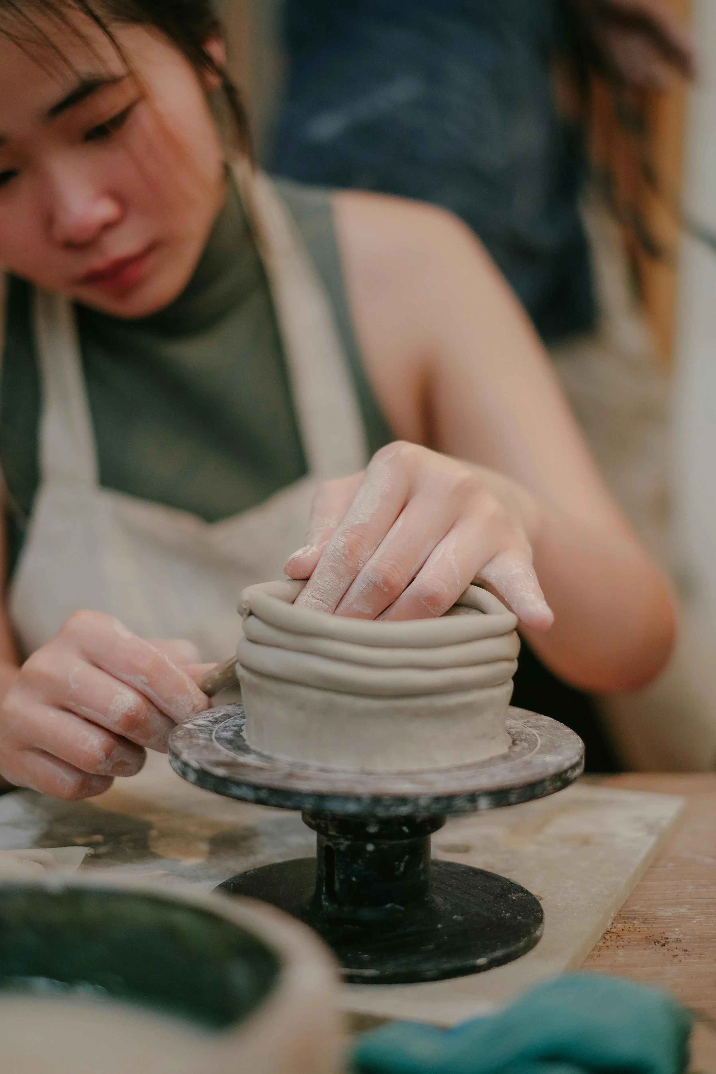 A person shaping a clay pot on a pottery wheel, with hands covered in clay, in a ceramics studio.