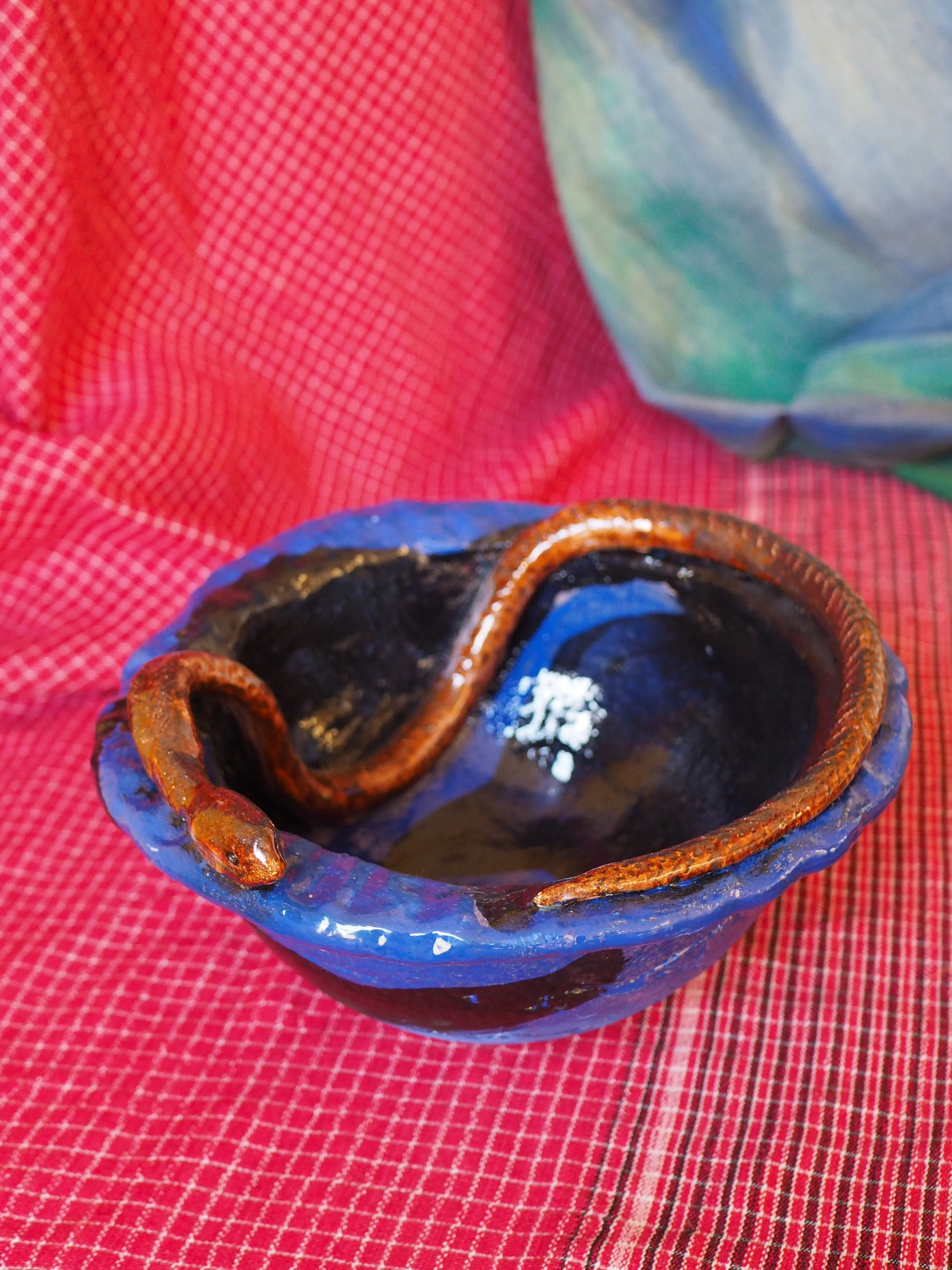A small, rustic pottery cup with a dark interior and a brown, textured handle, sitting on a red checkered cloth.