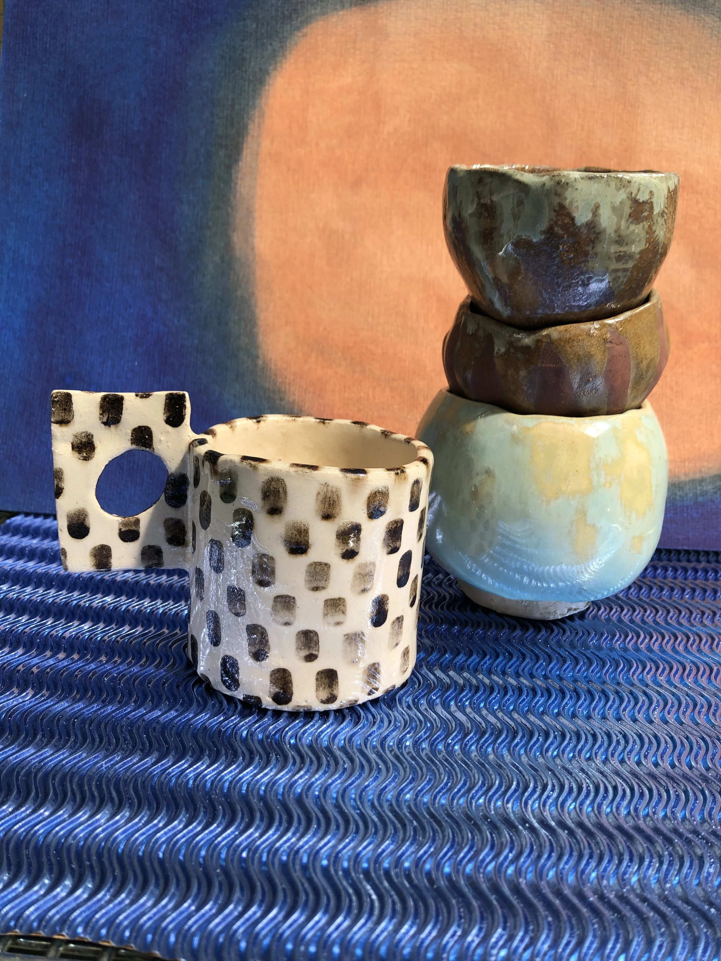 A collection of ceramic cups and bowls with various patterns, including some with polka dots and others with a marbled or glazed finish, placed on a textured blue fabric surface with a colorful gradient background.