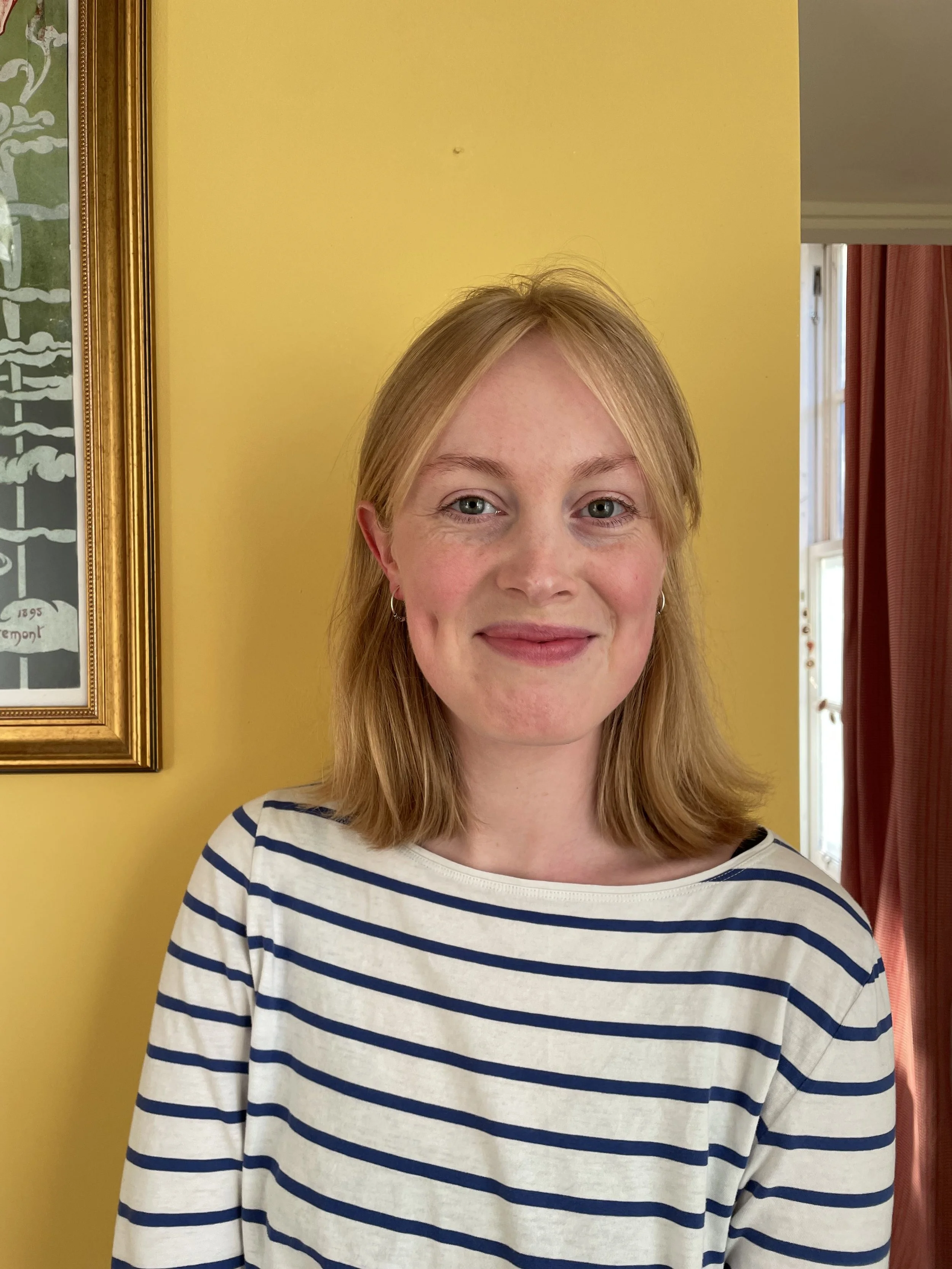 Lily Hannigan, a woman with blonde hair wearing a white and navy striped shirt, smiling, standing in front of a yellow wall with a framed picture and window with red curtains.