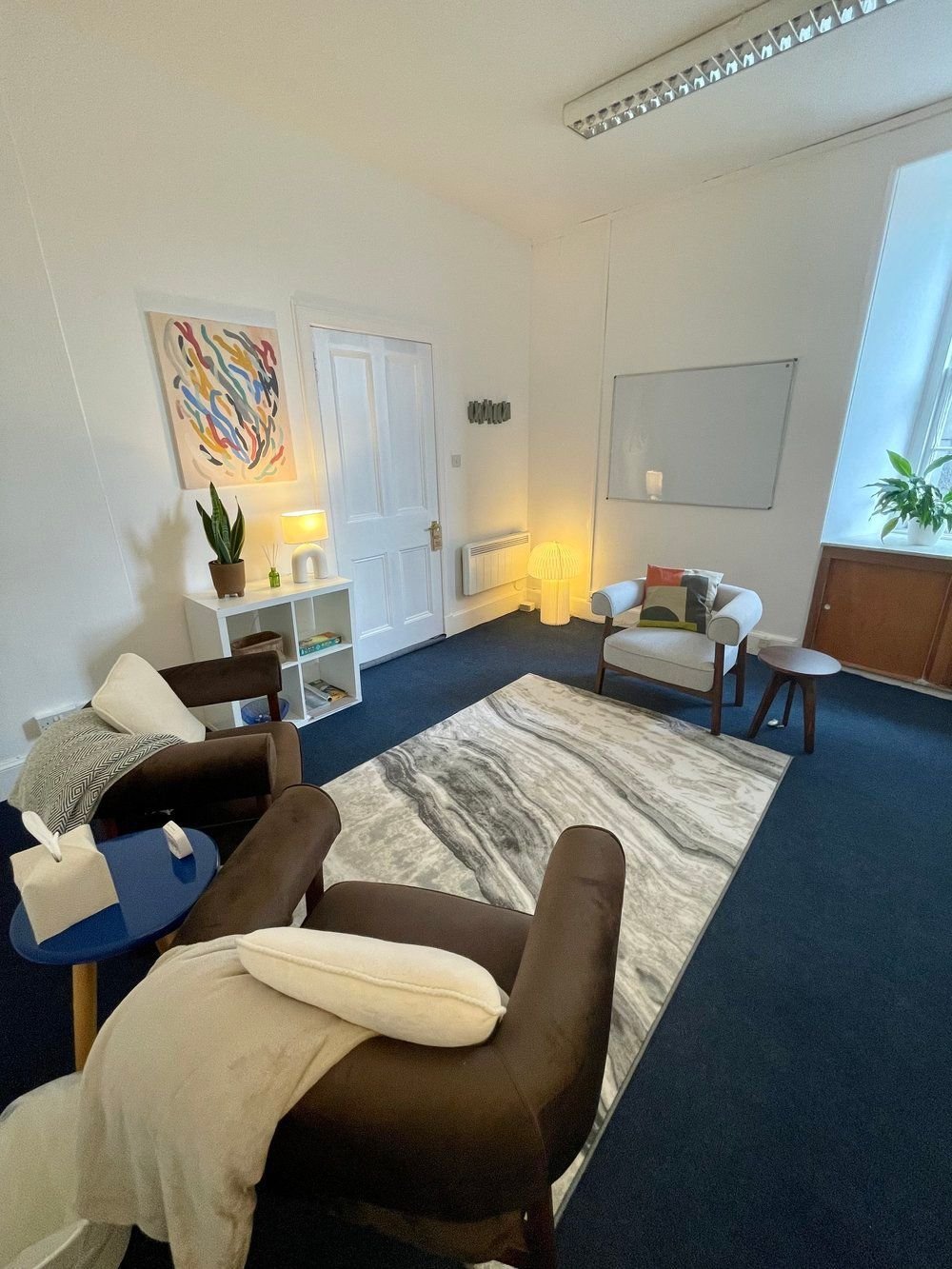 Living room with modern decor, featuring a white wall, a door, a white bookshelf with plants and books, a brown armchair with a white pillow and beige blanket, a small blue table with tissues, an abstract artwork, a patterned rug, a white armchair with colorful cushions, a small round table, a floor lamp, window with plants, and a whiteboard on the wall.