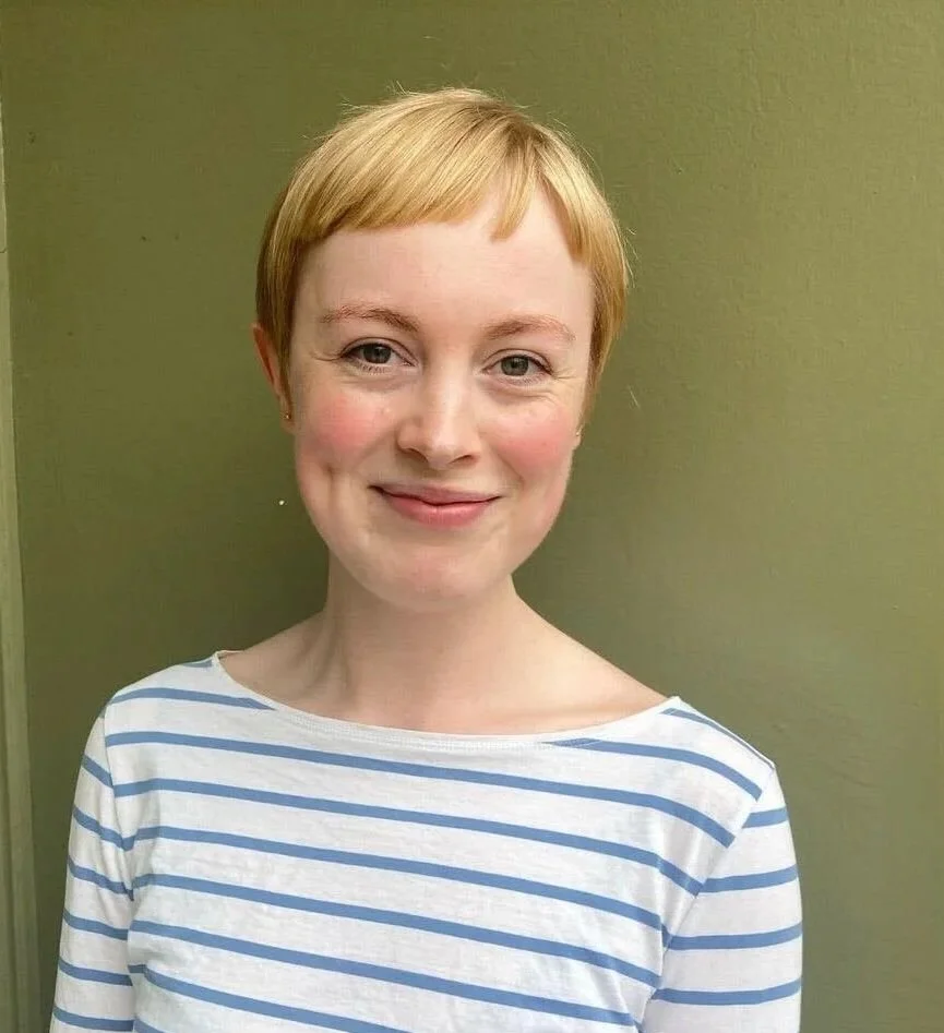 A woman with short blonde hair, fair skin, and a friendly smile, wearing a white and blue striped shirt, standing in front of a plain olive-green wall.