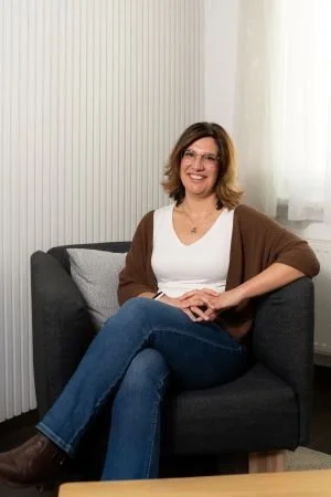 Babsi sitzt auf einem grauen Polstersessel  in einem Raum mit weißen Wänden und einem Fenster mit weißen Vorhängen, sie lächelt und trägt eine weiße Bluse und eine braune Strickweste.