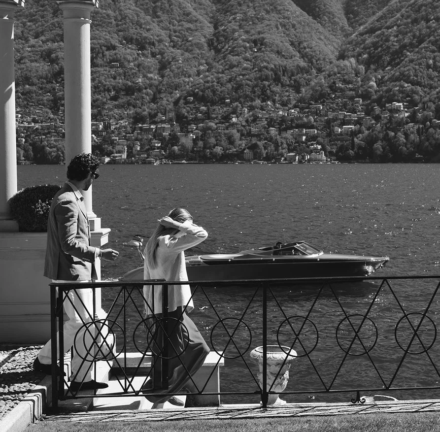 Two people walking by a waterfront with a boat, hills, and houses in the background