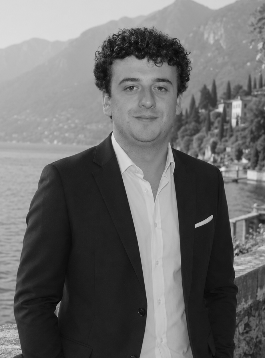 A young man with curly hair in a suit, standing outdoors near a lake with mountains and buildings in the background.