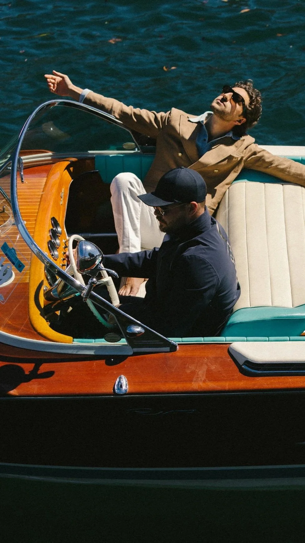 Two men in a vintage wooden boat on water, one driving and the other relaxing with arms outstretched, wearing sunglasses and casual clothing.
