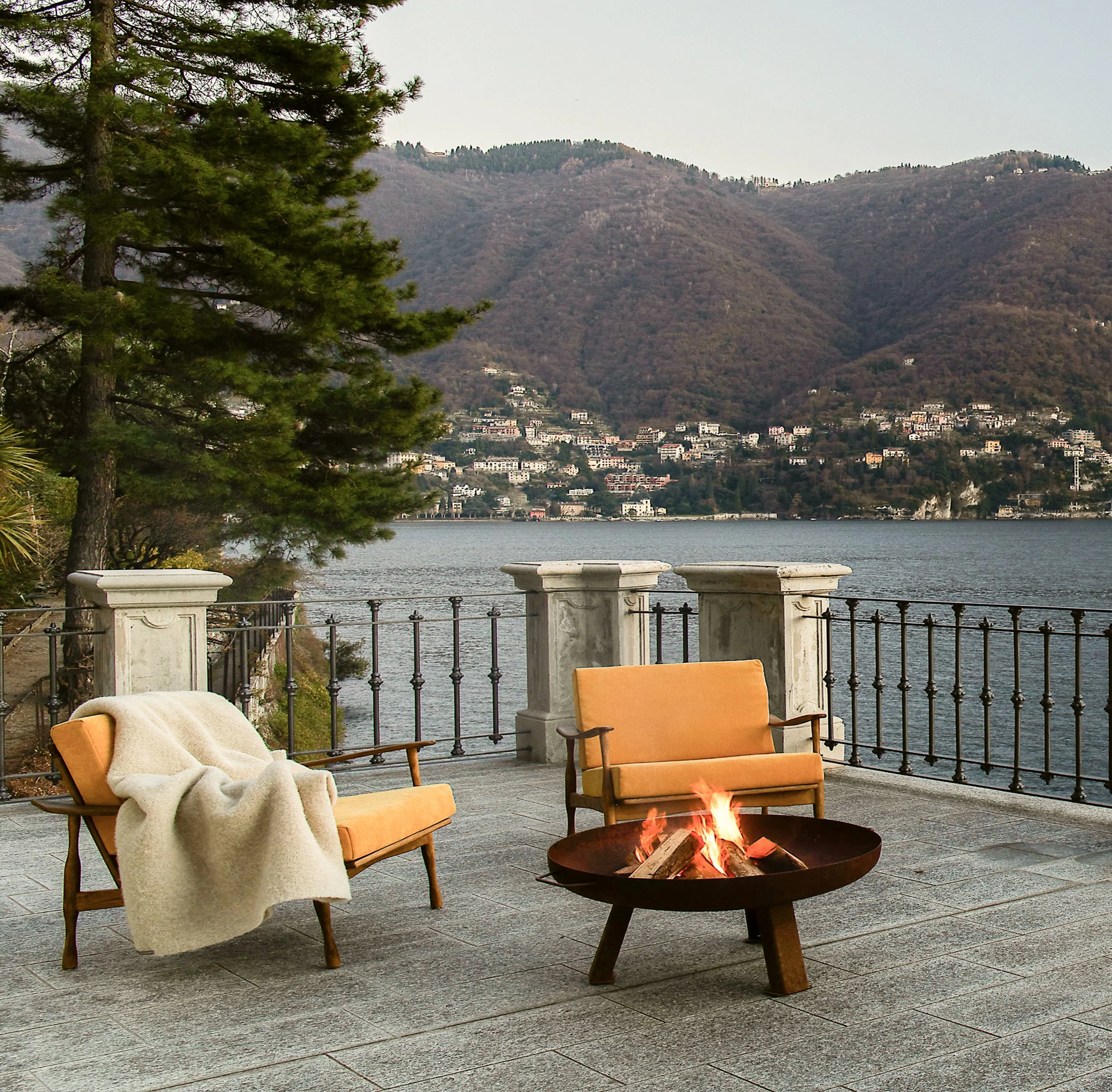 Outdoor patio area with two orange chairs and a fire pit, overlooking a body of water and hillside with buildings and trees in the background.