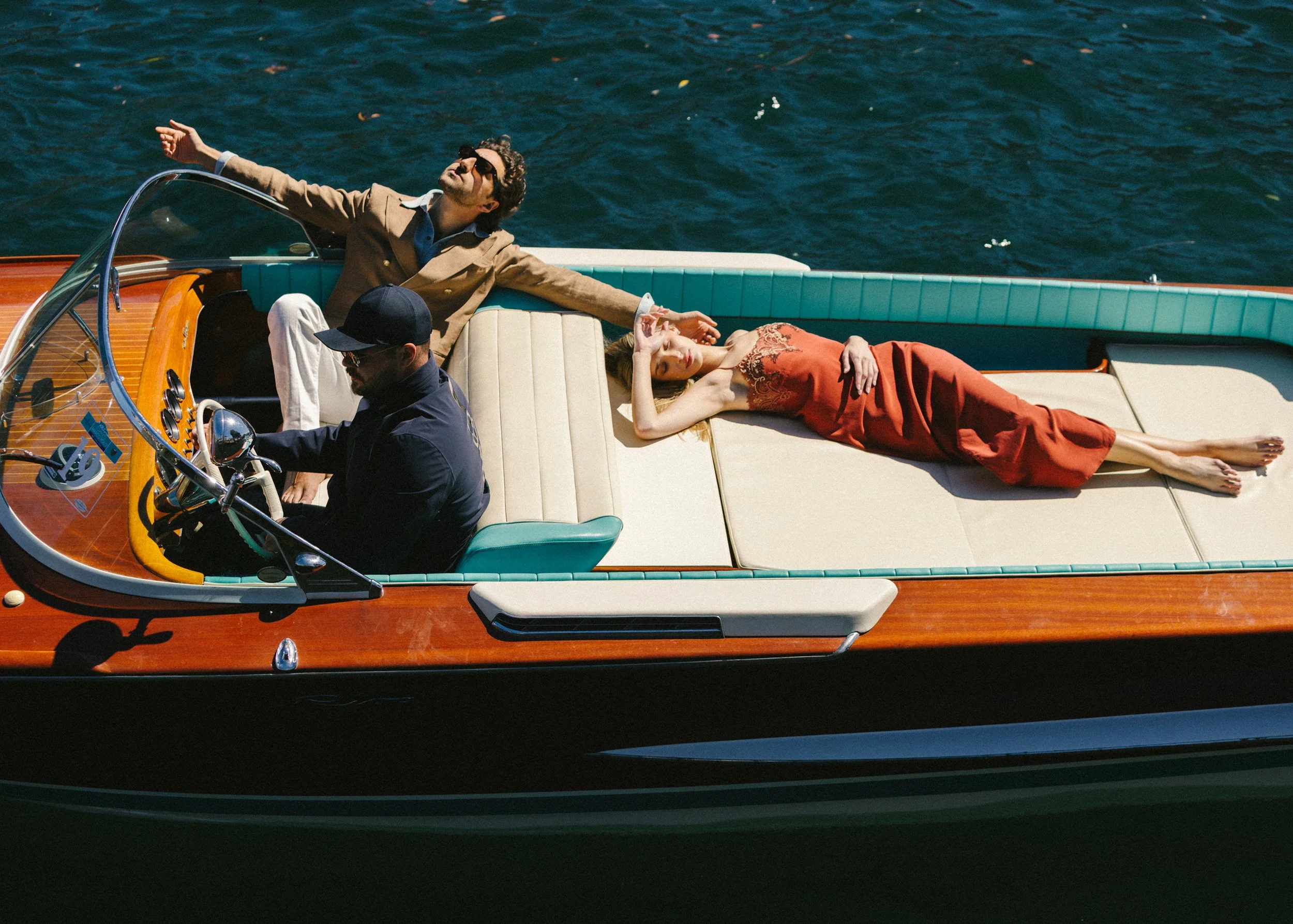 A man and a woman relaxing on a boat with a man driving, water in the background.