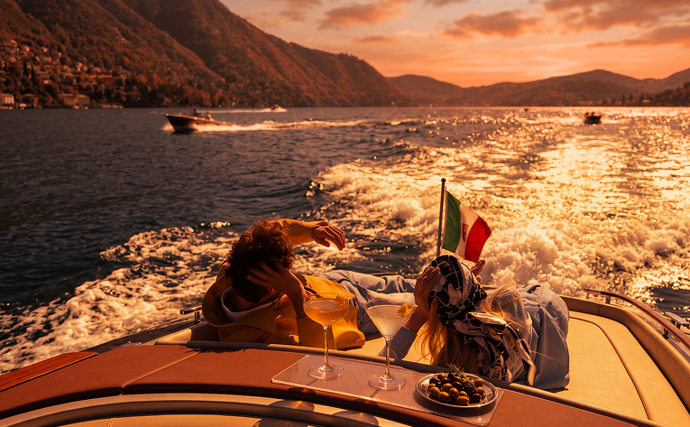 Two women are relaxing on a boat during sunset, with one holding an Italian flag. They have drinks and a small bowl of olives on the boat's surface, with water and other boats visible in the background.