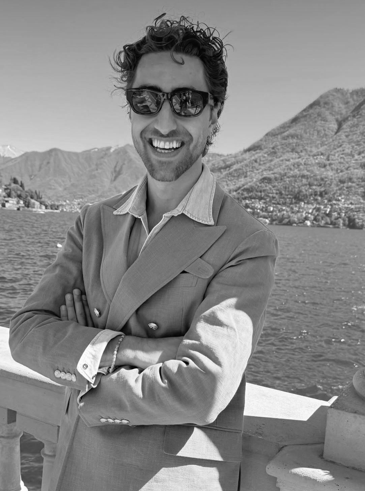 A man in sunglasses and a light-colored double-breasted blazer smiling with arms crossed, standing outdoors near a body of water with mountains and a small town in the background.