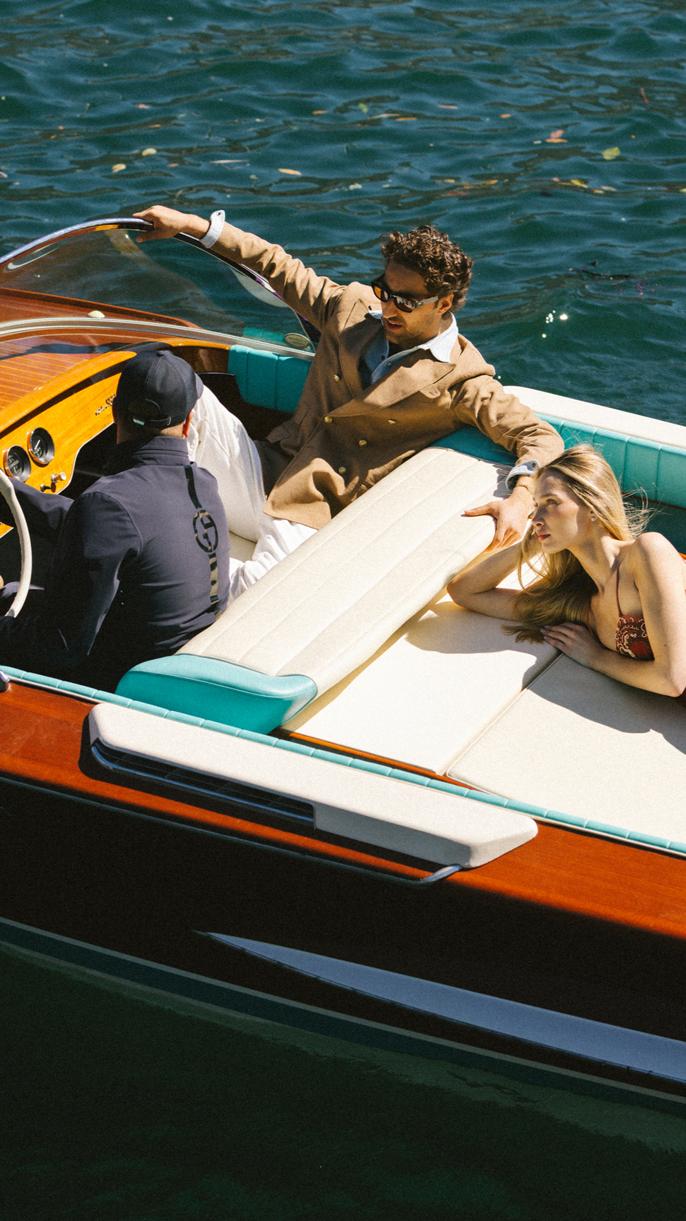 A man in sunglasses and a brown jacket sitting on a boat with a woman lying next to him in a bikini, all on a body of water.
