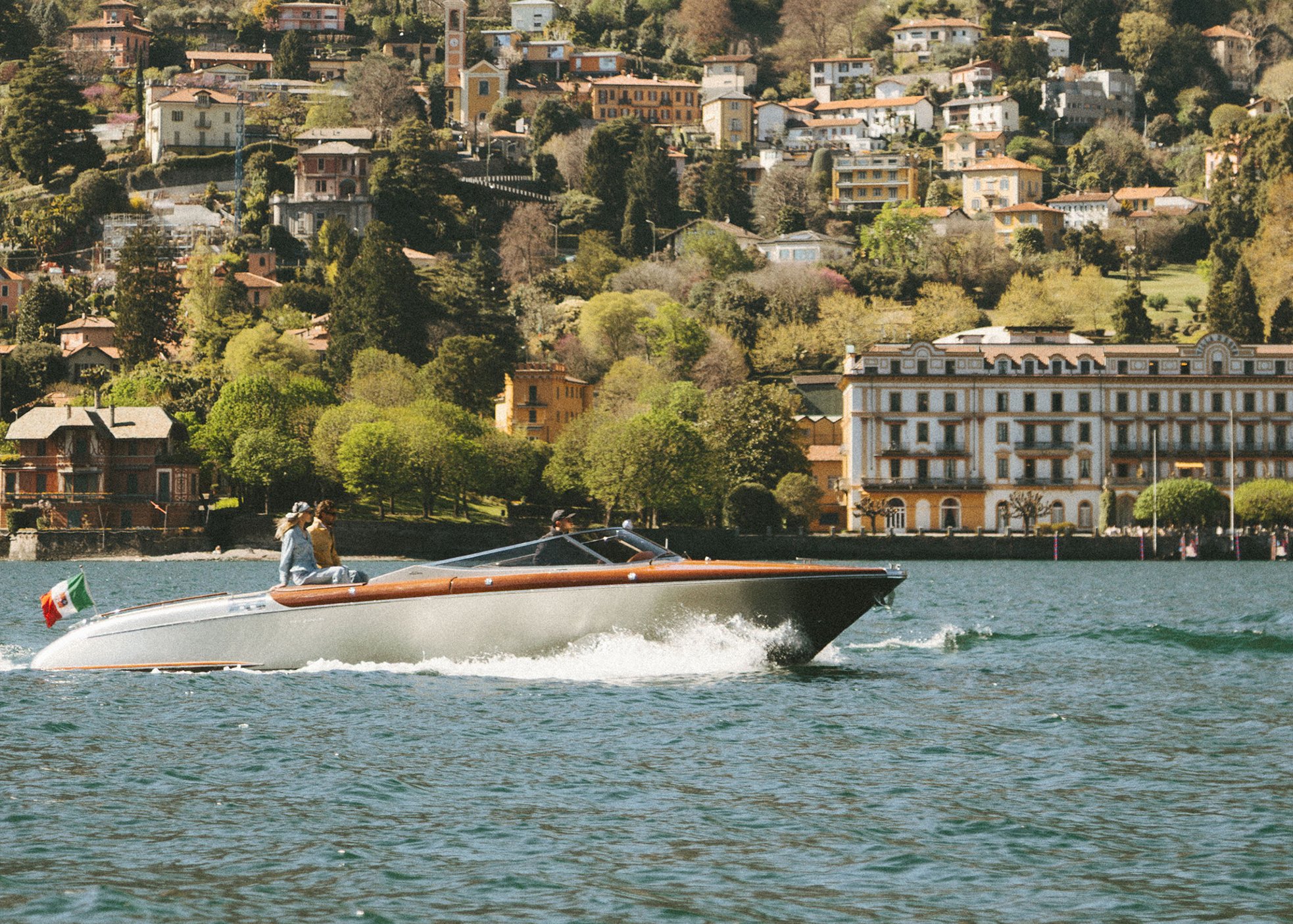 People enjoying a boat ride on a lake with a hilly residential area in the background, featuring colorful houses and lush greenery.