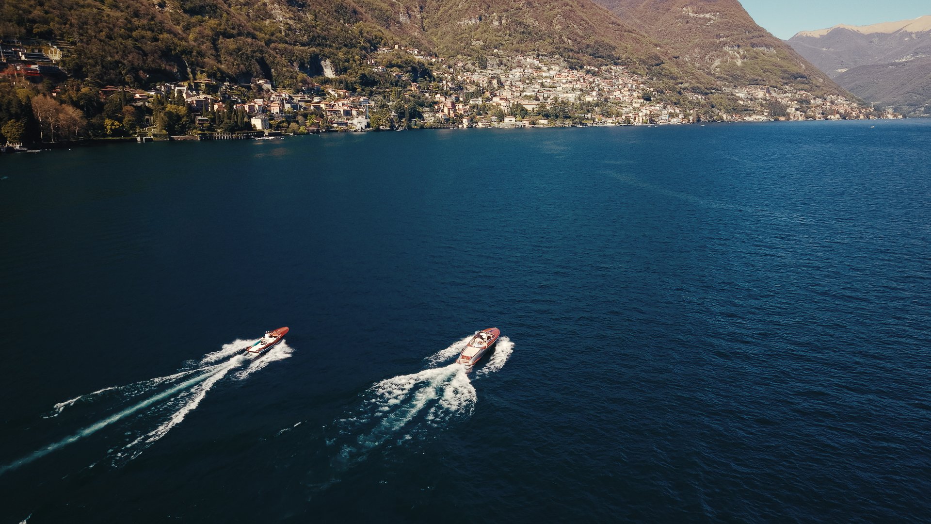 Two boats speed across a large body of water near a mountainous shoreline with residential buildings.