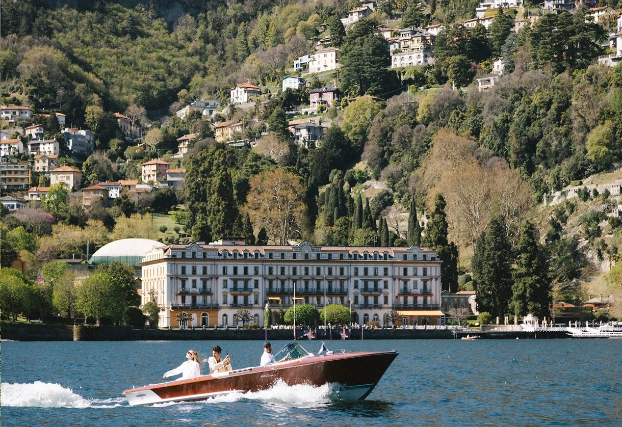 A boat with three people on a lake, with a hillside residence and lush greenery in the background.