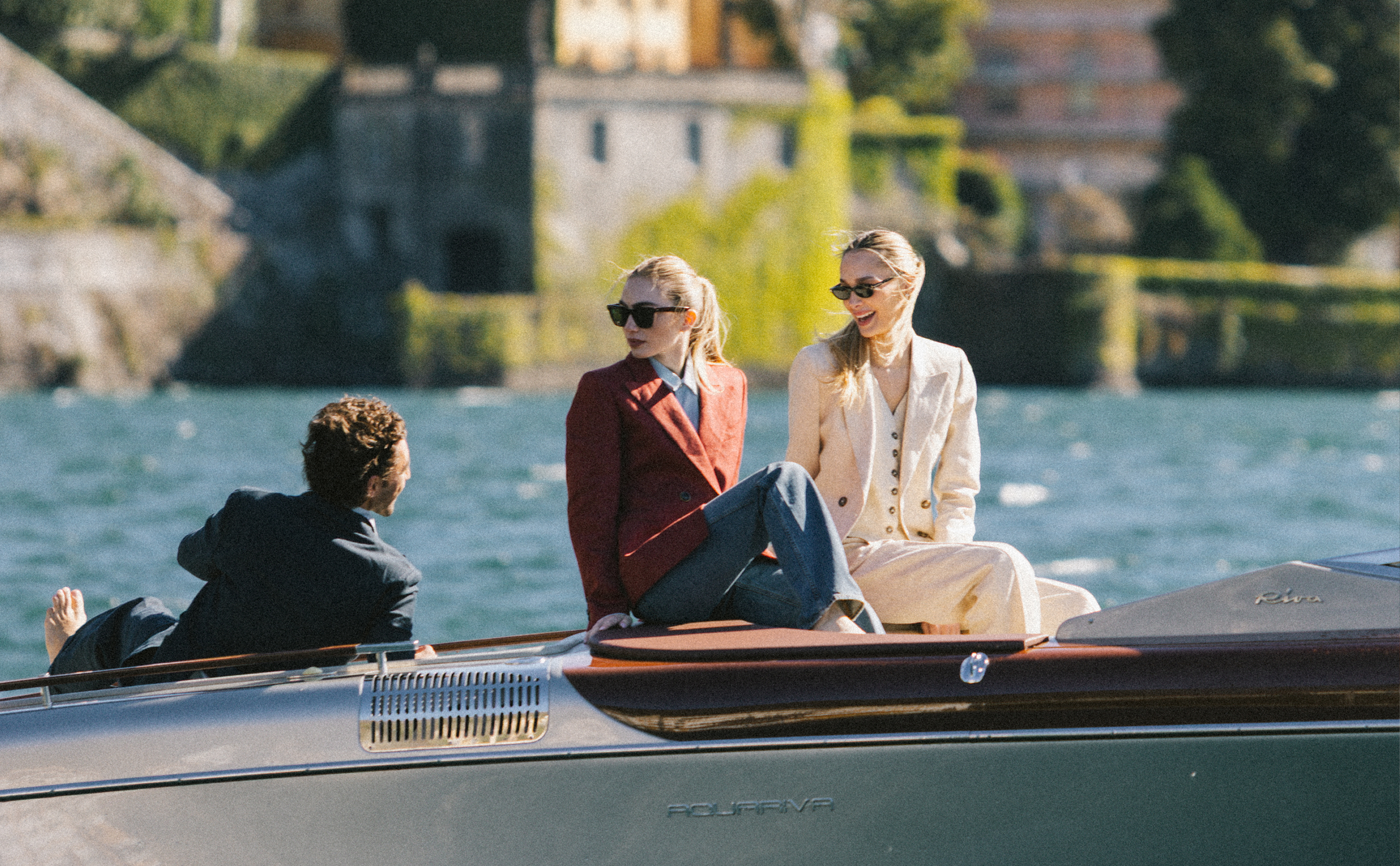 Three women on a sailboat, two standing and one lying down, enjoying a sunny day on the water with a castle-like building and trees in the background.