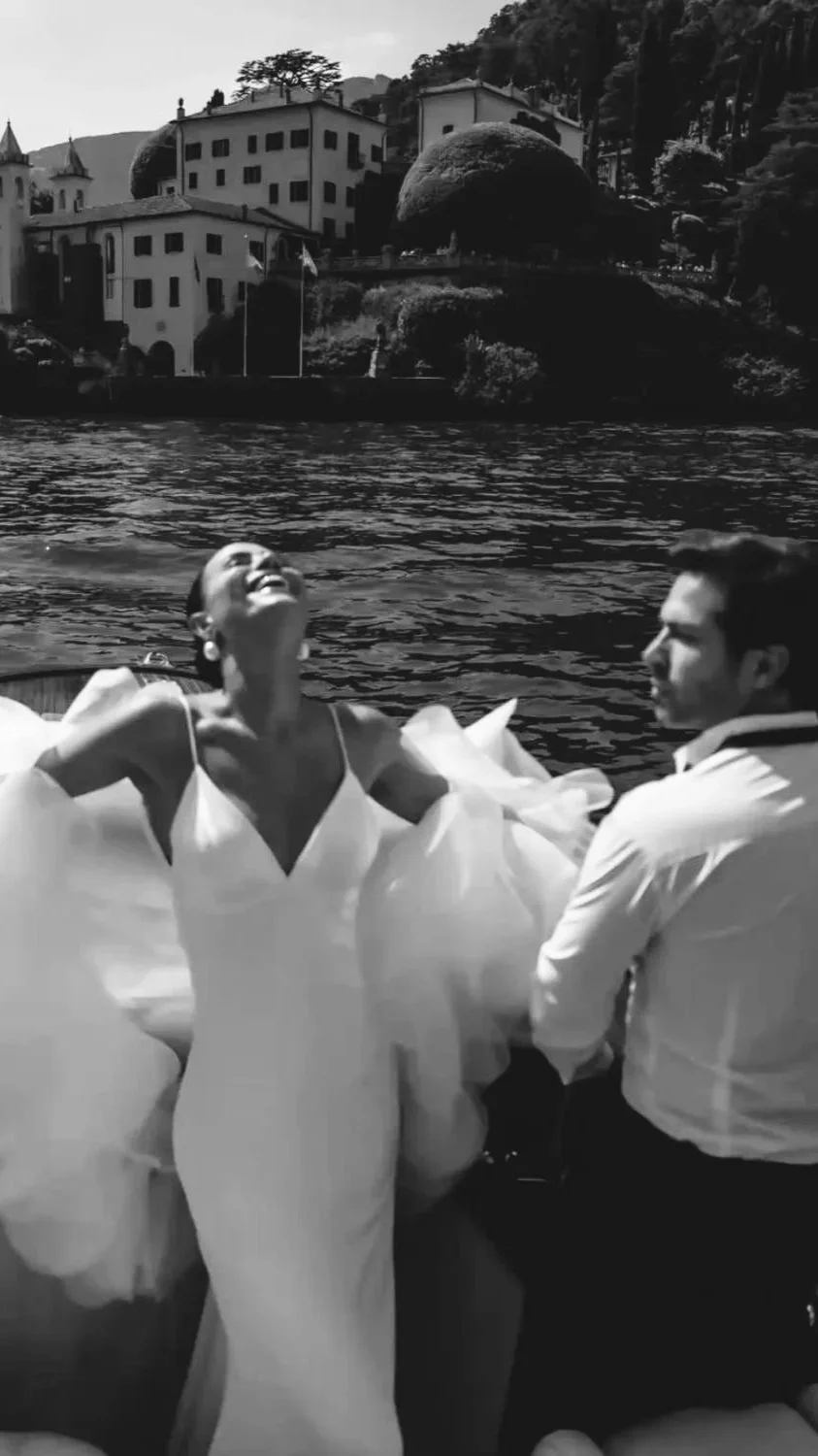 A woman in a flowing white dress laughing happily while sitting on a boat with a man next to her by a lake with houses and trees in the background.