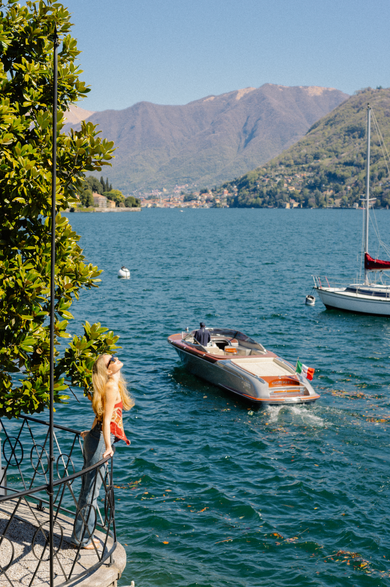 A woman with long blonde hair and sunglasses stands on a balcony overlooking a lake, with boats on the water, surrounded by mountains.