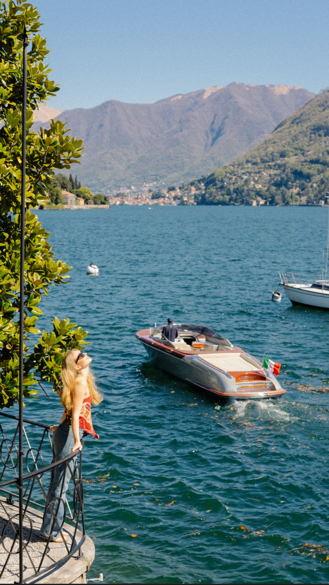 A woman with blonde hair and sunglasses stands on a balcony by a lake, watching a speedboat with an Italian flag move across the water. The lake is surrounded by mountains and small houses.