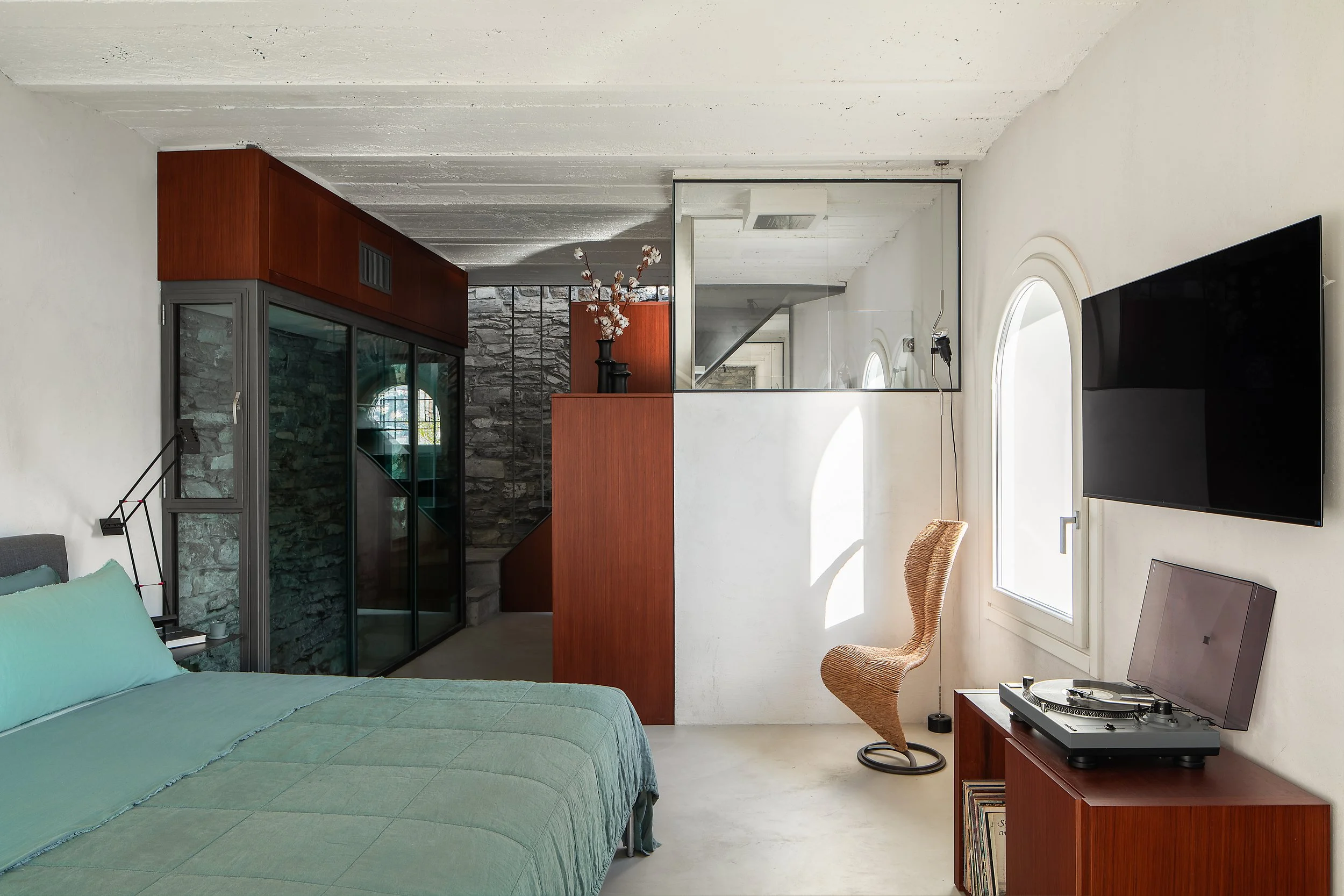 A modern living room with a bed, a TV on a wooden stand, and a turntable. The room features a glass partition, a decorative vase with flowers, and curved windows.