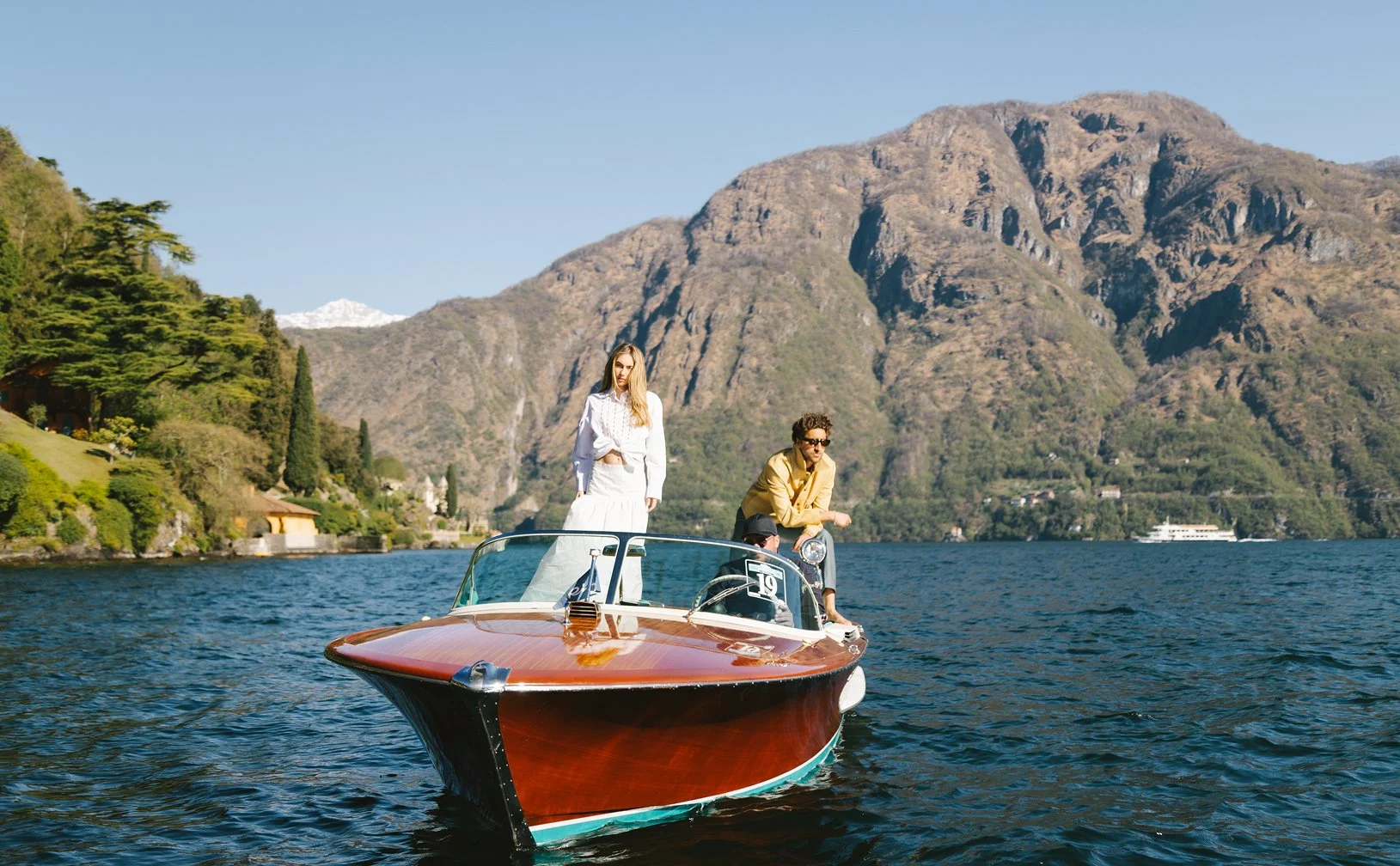 Two women and a man on a wooden speedboat on a lake, with mountains and trees in the background on a clear day.