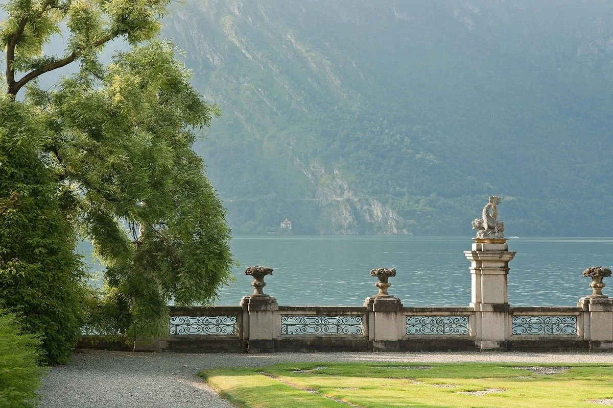 A scenic lakeside view with a stone decorative railing and a statue of a dragon on a pedestal, surrounded by lush greenery and mountains in the background.
