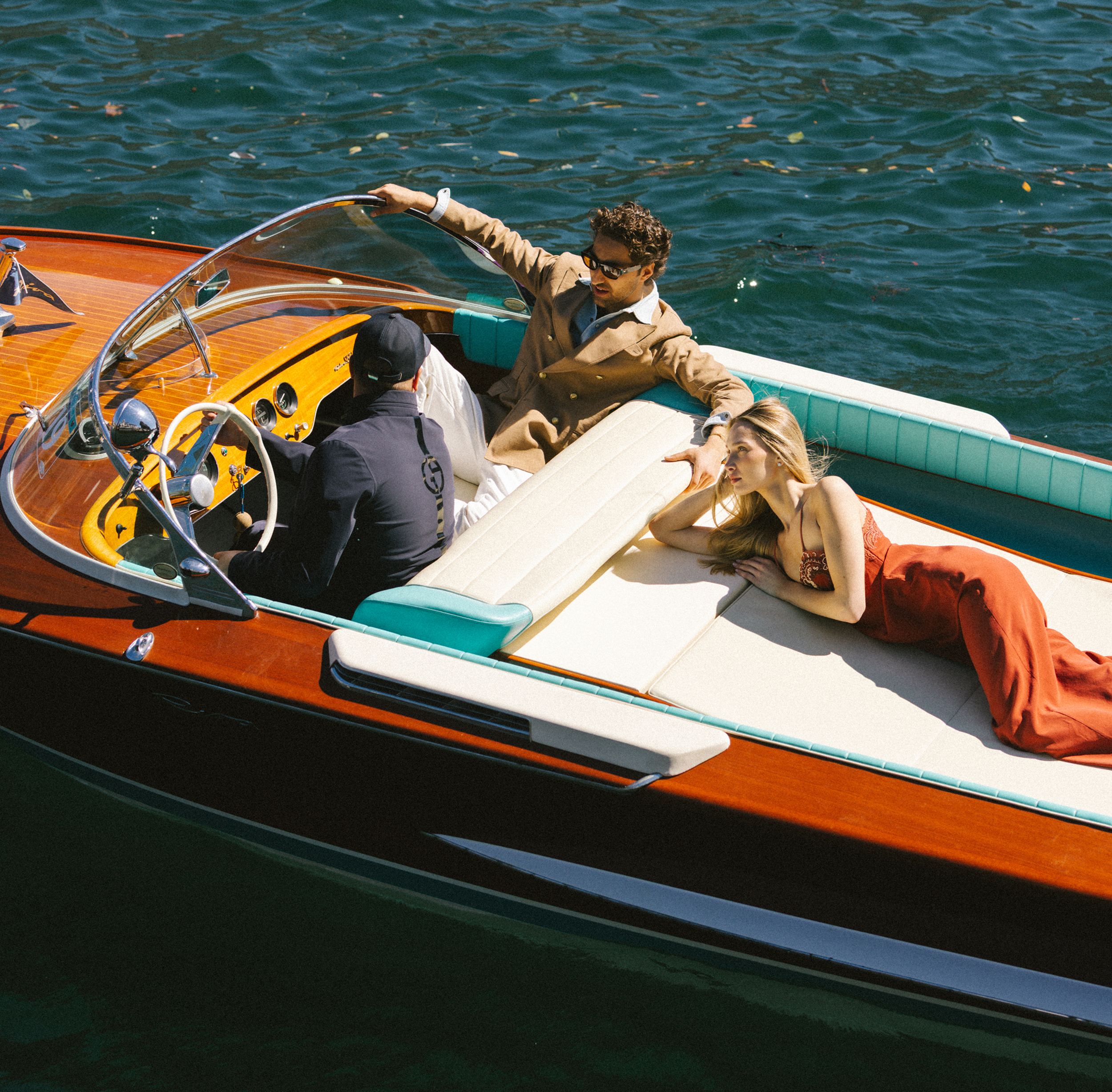 A group of four people on a wooden and white boat on the water. One man with curly hair and sunglasses is gesturing while talking to a woman lying on the boat's edge, and another person is sitting with their back to the camera.
