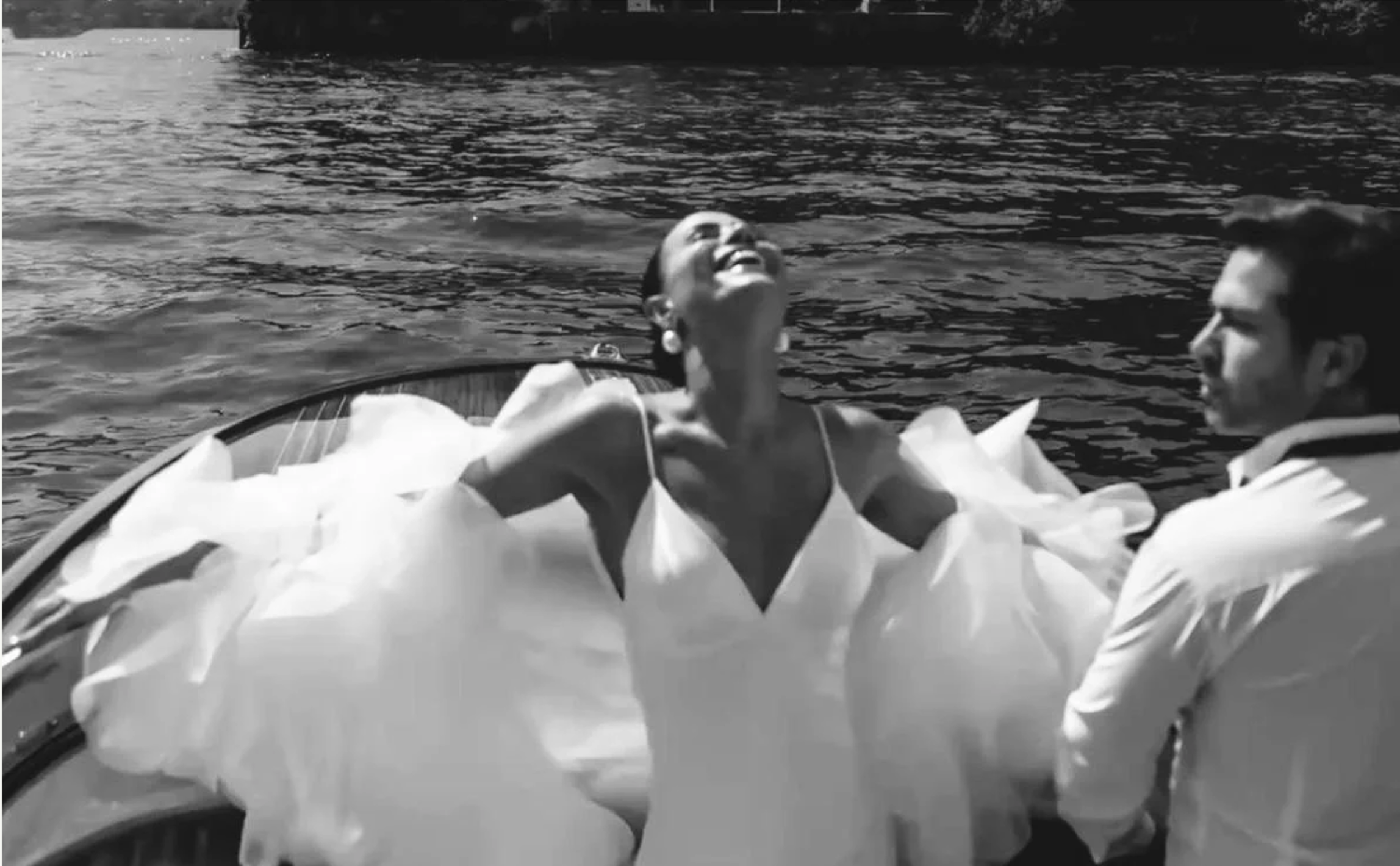 A woman is smiling, leaning back with her head tilted upwards, on a boat with another individual partially visible. The scene is on a body of water with a dock or structure in the background. The image is in black and white.
