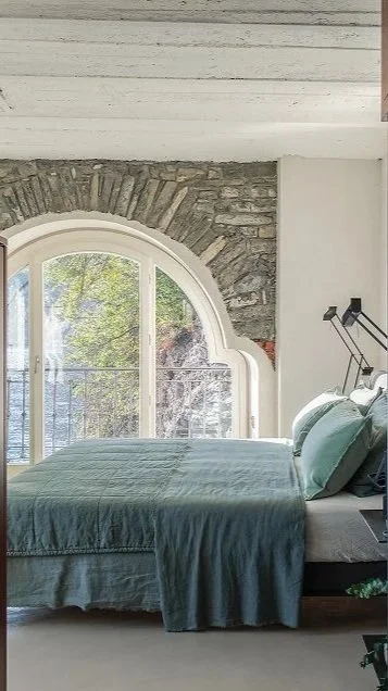 A modern bedroom with a large arched window, stone wall accents, a neatly made bed with green bedding and pillows, and two black adjustable wall-mounted reading lamps.