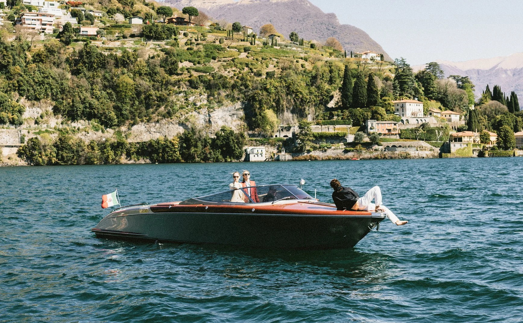 A luxury motorboat on a lake with three people on board, two women and one man. The women wear sunglasses and light-colored clothing, while the man is sitting on the bow with his legs dangling in the water. The background shows a hillside with lush greenery and houses along the shoreline.