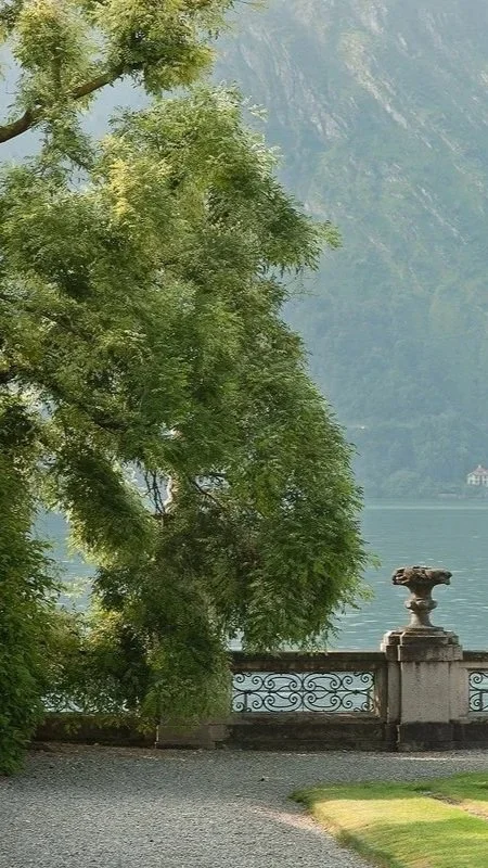 A lakeside scene with green trees, a stone railing, and a small sculpture on a pedestal, with mountains and water in the background.