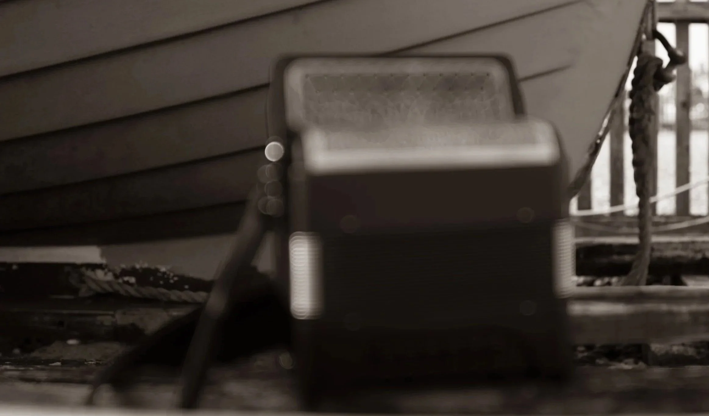 A blurry black and white photo of a bench on a porch or deck with horizontal siding in the background, a chain securing the bench, and a couple of logs on the ground.