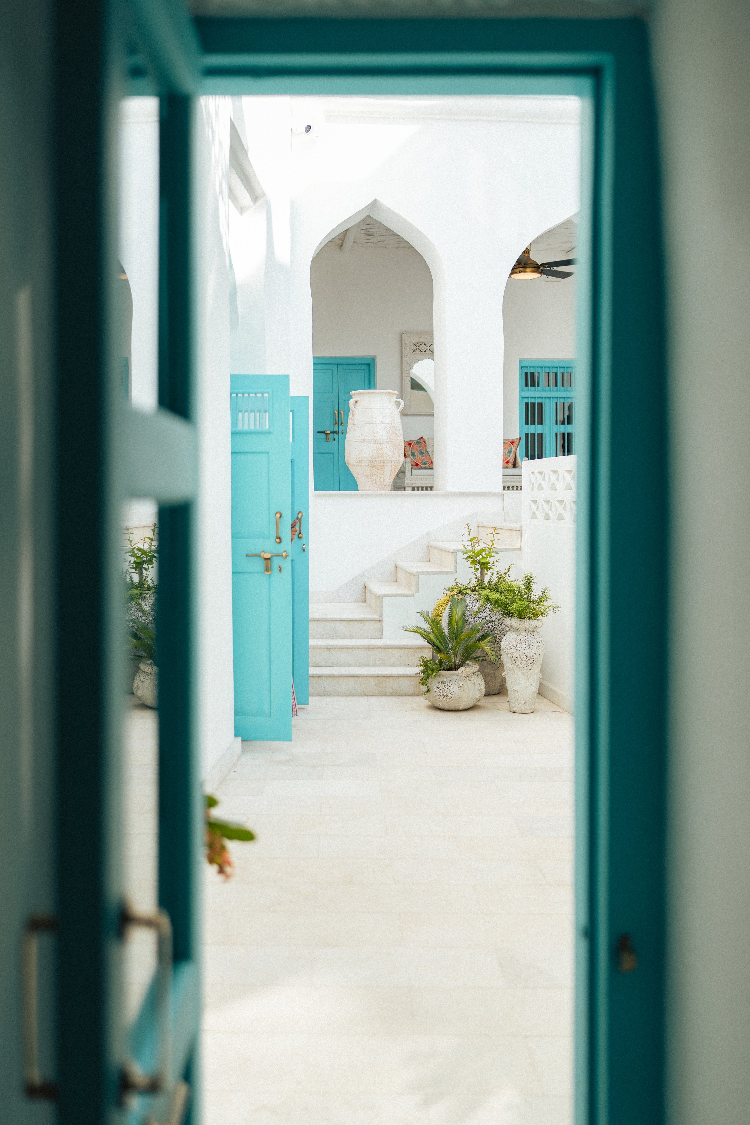 View through a window showing a white courtyard with arched openings, blue doors, white steps, potted plants, and outdoor seating with colorful cushions.