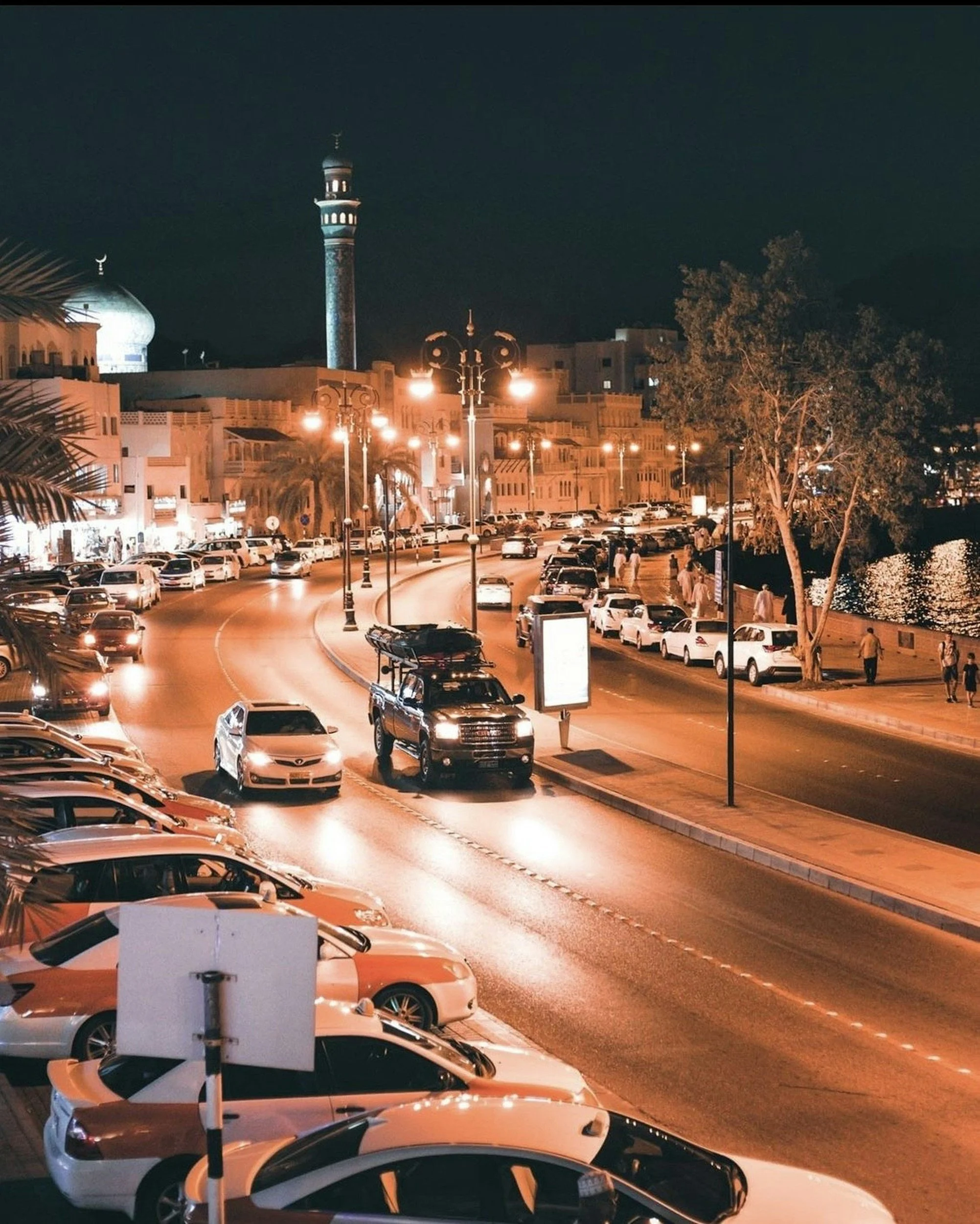 A night scene of a street with parked cars, streetlights, a tree, a mosque with a blue dome, and a minaret in the background
