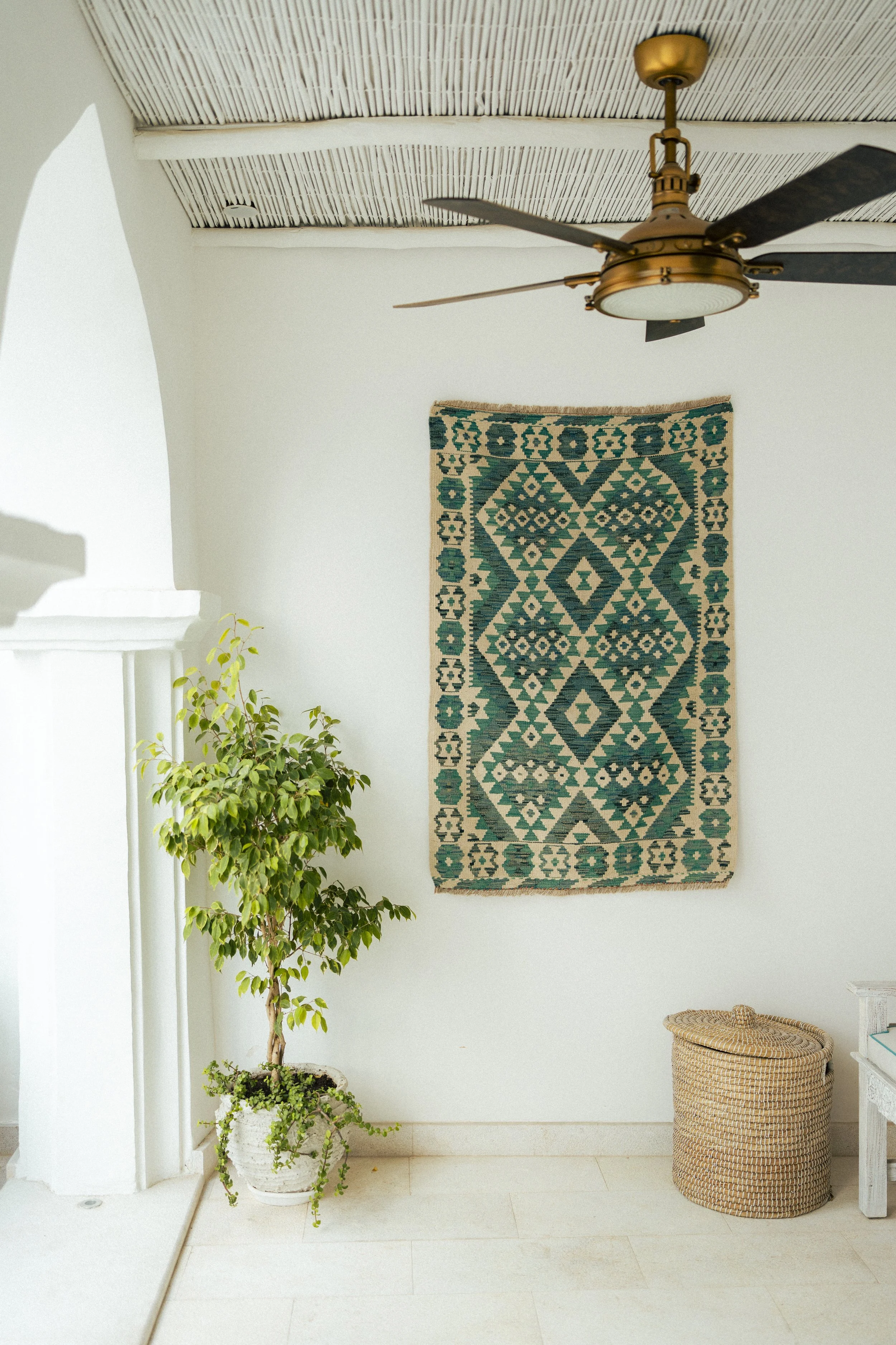 Interior of a room featuring a hanging ceiling fan, a decorative rug on the wall with geometric patterns, a potted plant with green leaves, and a round wicker basket on the floor next to a small side table.