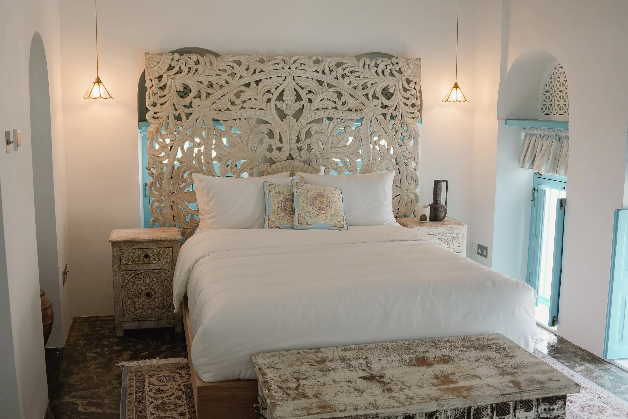 A bedroom with a white bed, floral patterned pillows, and an ornate carved wooden headboard. There are two hanging pendant lights, a bedside table with decorative items, and a small window with ruffled curtains.