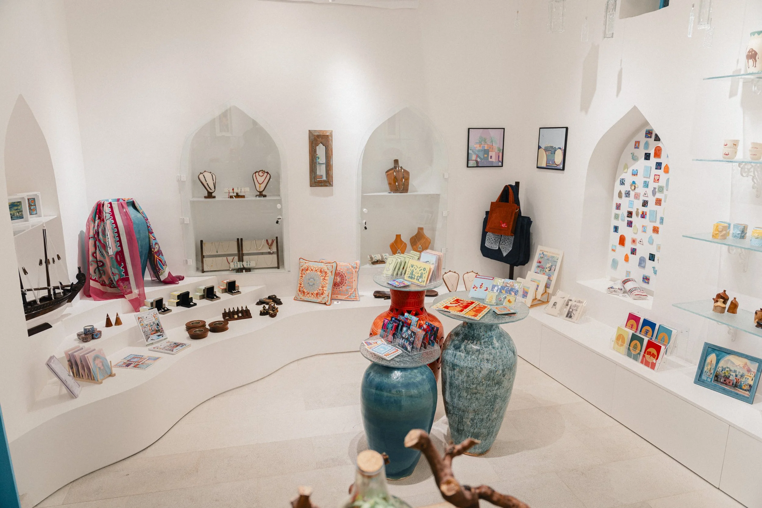 Interior of a Mezrad store displaying jewelry, artwork, and decorative items on white shelves and tables, with blue vases in the foreground, against white walls with artwork.