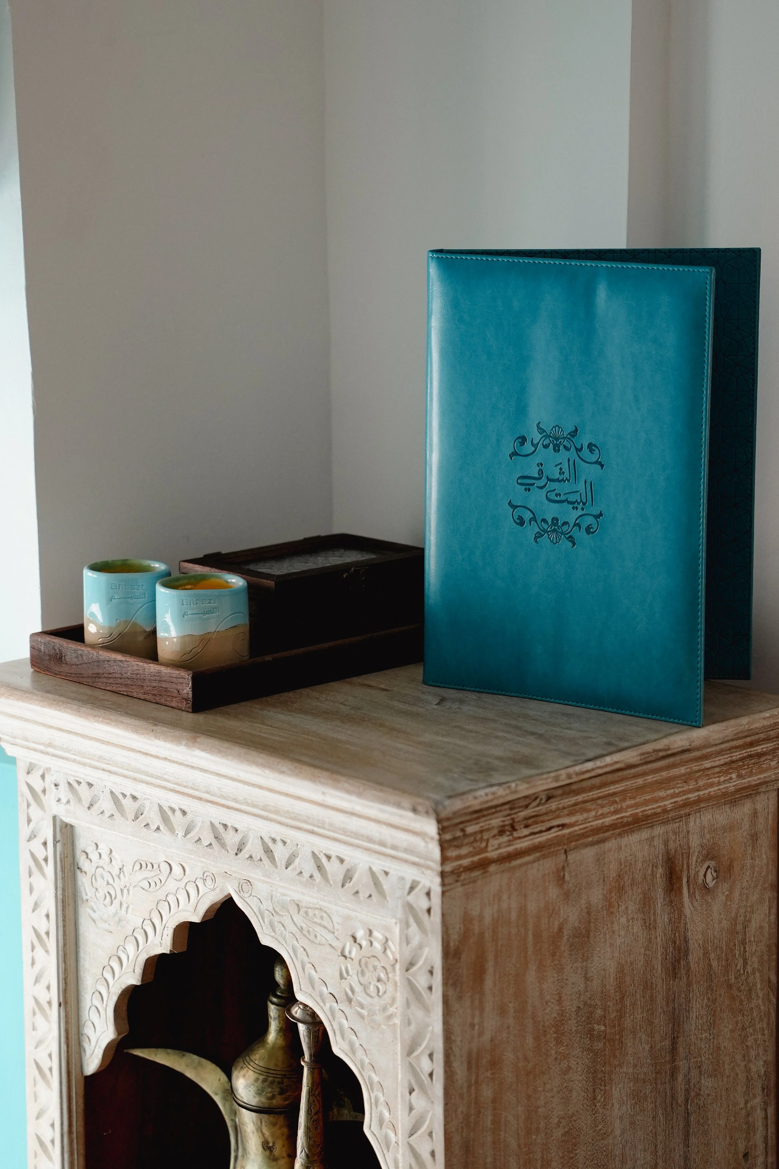 A wooden table with two small ceramic cups and a decorative wooden box. Behind the table, there's a blue menu or book with Arabic writing and floral designs. Part of a white carved wooden structure is visible at the bottom of the image.