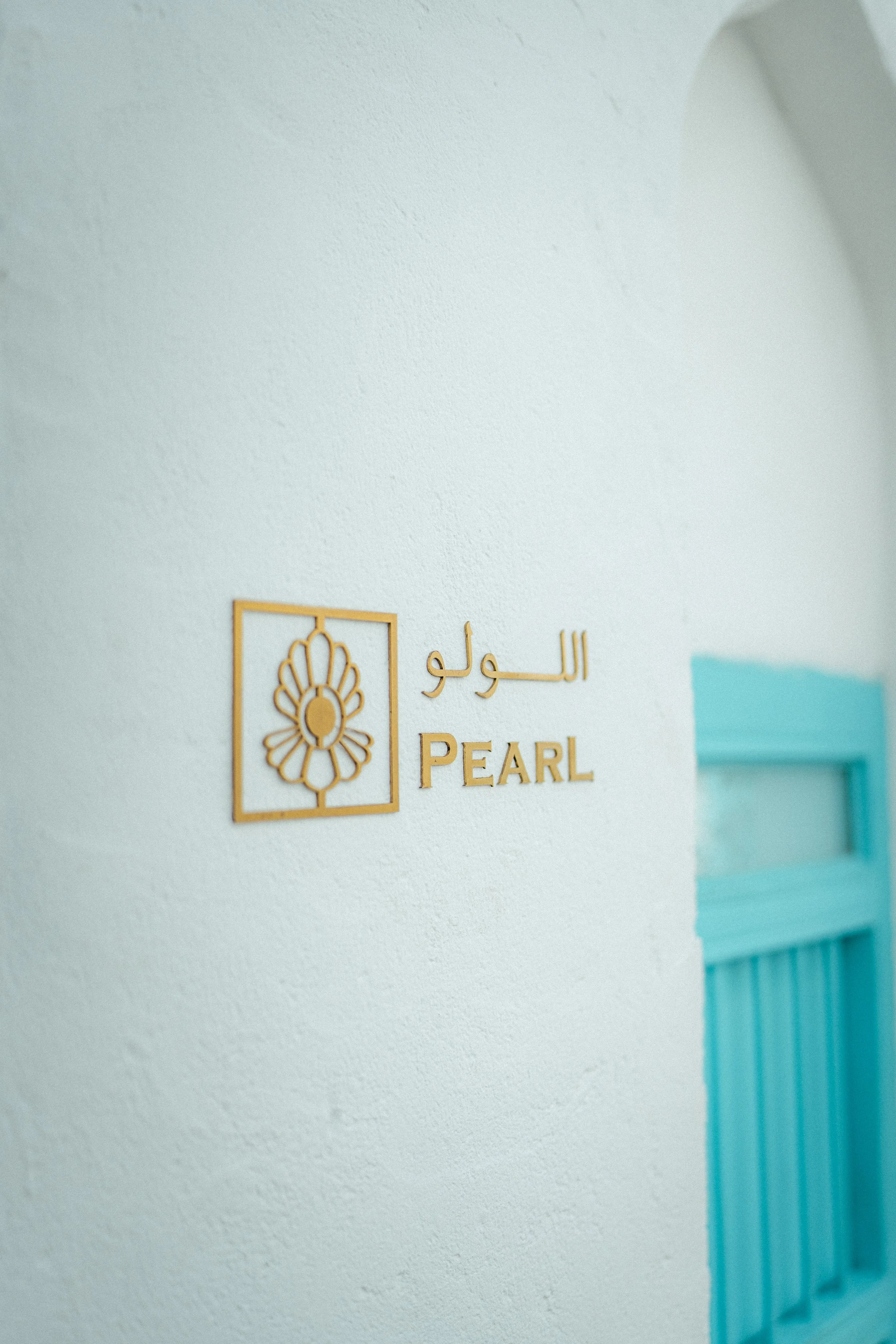 A wall with a decorative sign that reads 'PEARL' in English and Arabic, with a geometric floral design inside a square frame, and part of a turquoise door on the right side.