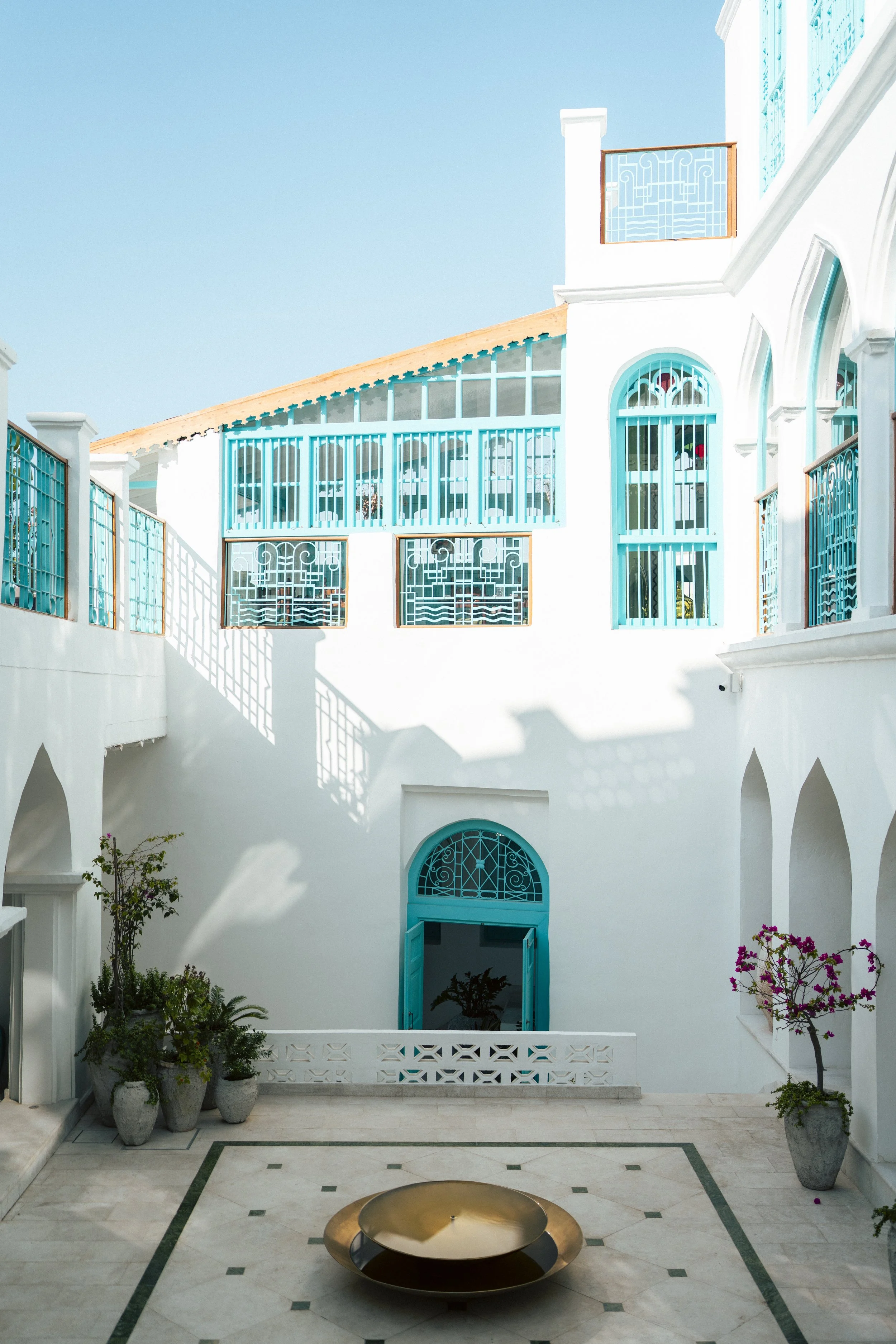 A white building with blue accents, including blue window frames, doors, and railings, features a courtyard with potted plants and a metallic fire pit in the center, under a clear blue sky.
