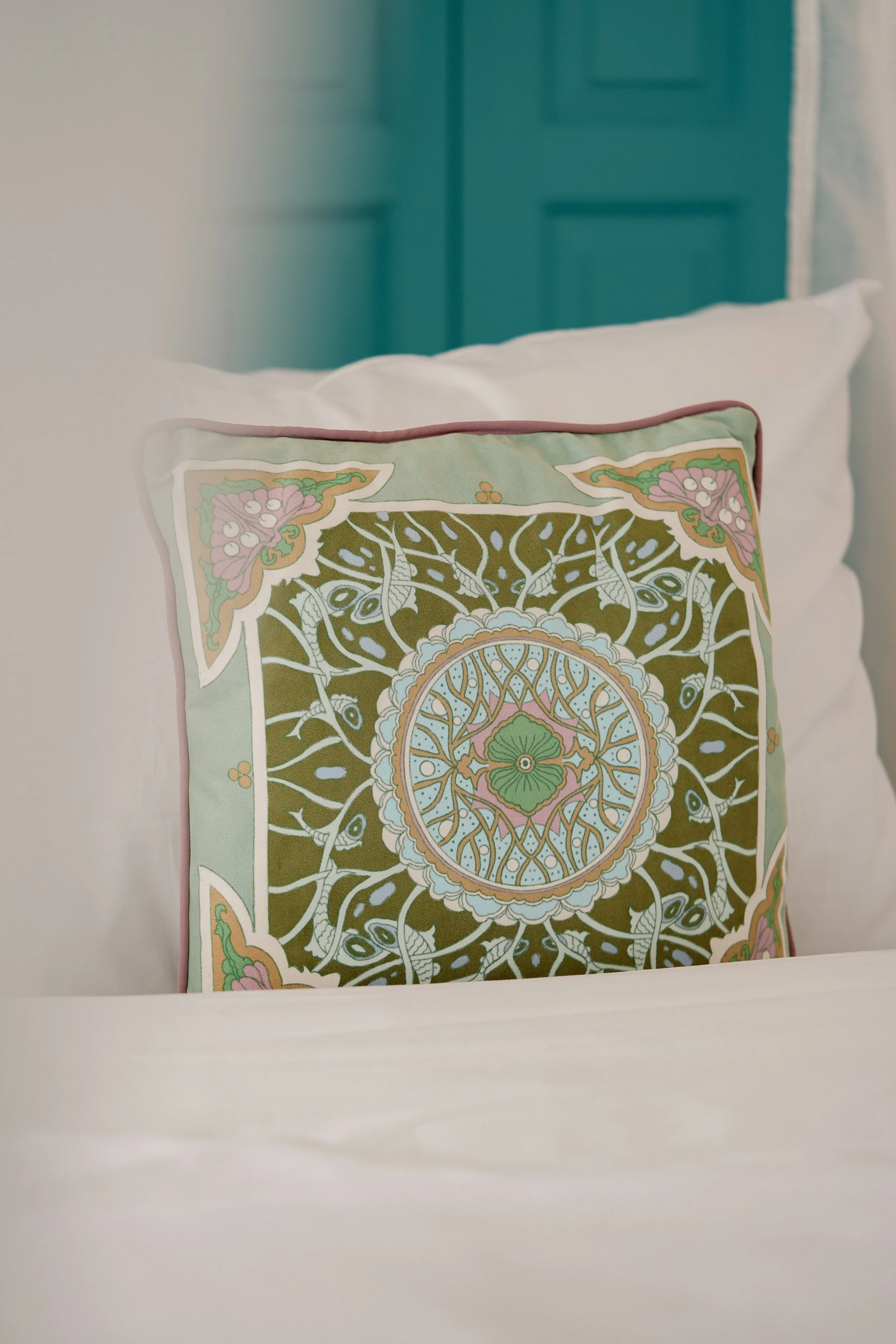 Decorative pillow with intricate floral and geometric patterns in pastel colors on a bed with white sheets inspired by the house.