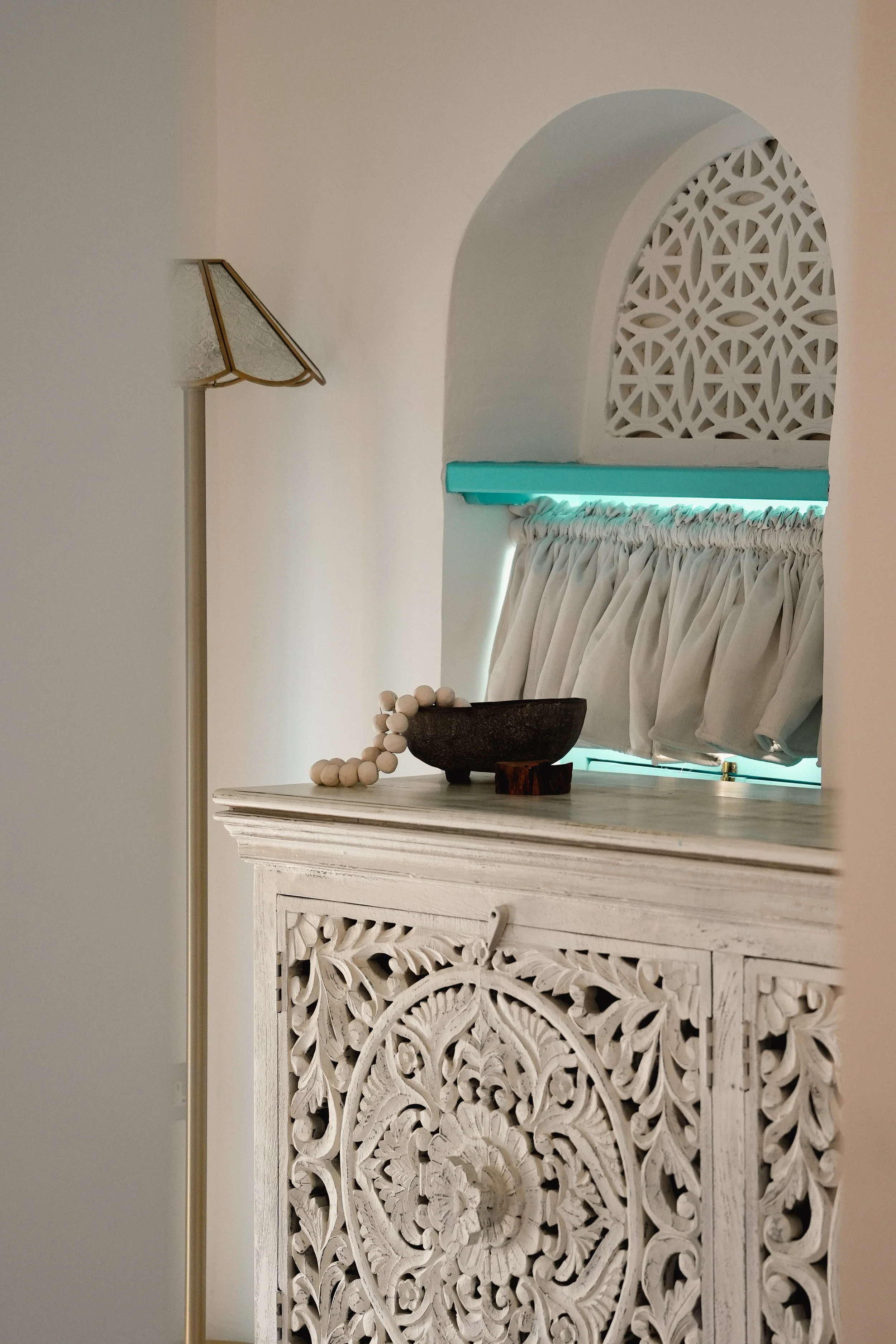 Interior room with white carved wooden furniture, a lamp with a beige shade, a decorative bowl, and a window with white curtains and an arched decorative screen.